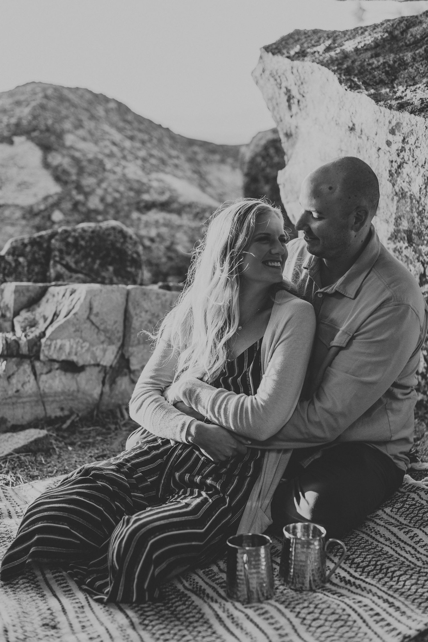 Black and white photo of couple cuddling on blanket against mountain backdrop.
