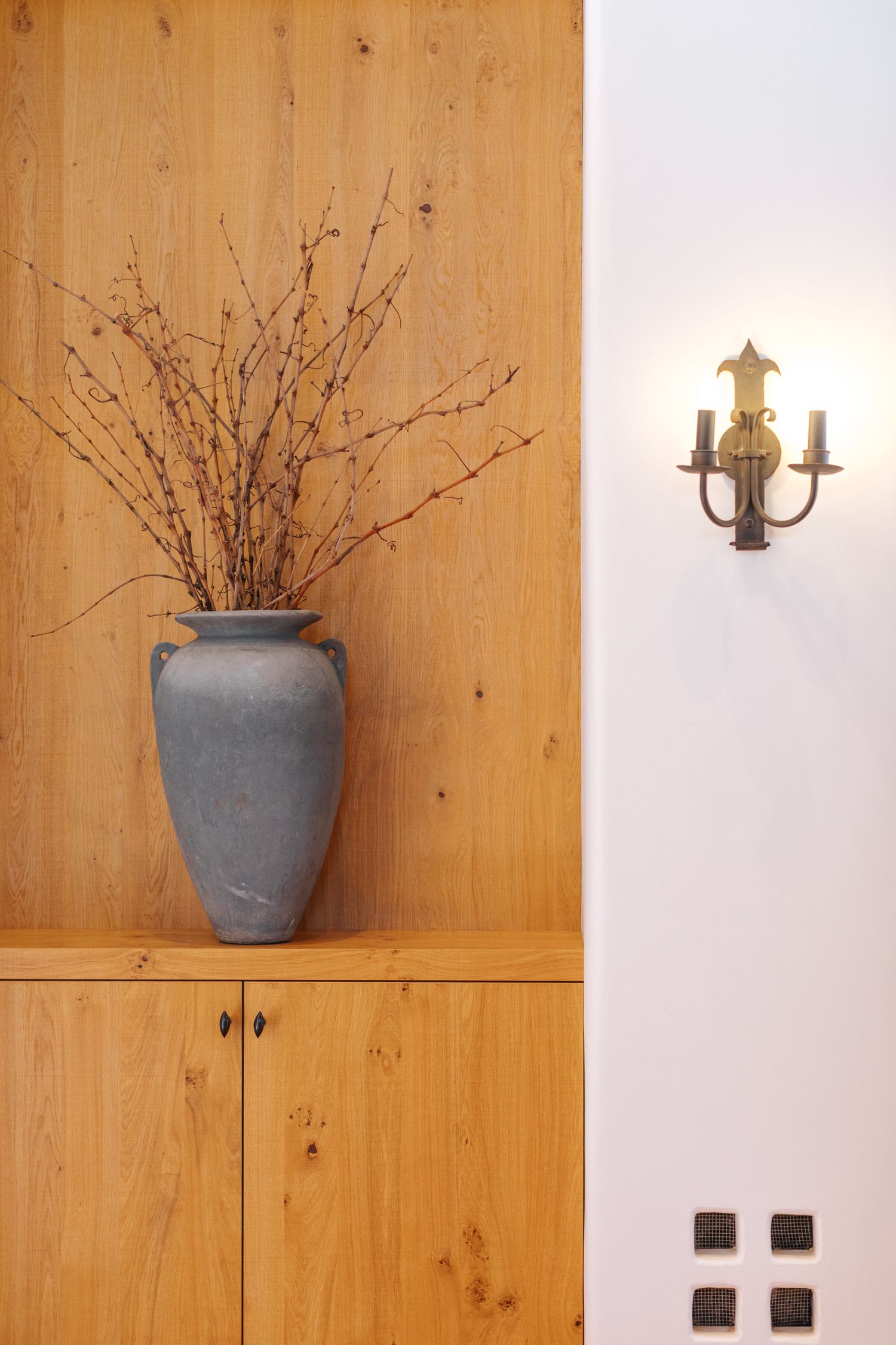 Wood-paneled wall with grey ceramic vase holding bare branches and wall sconce lighting.