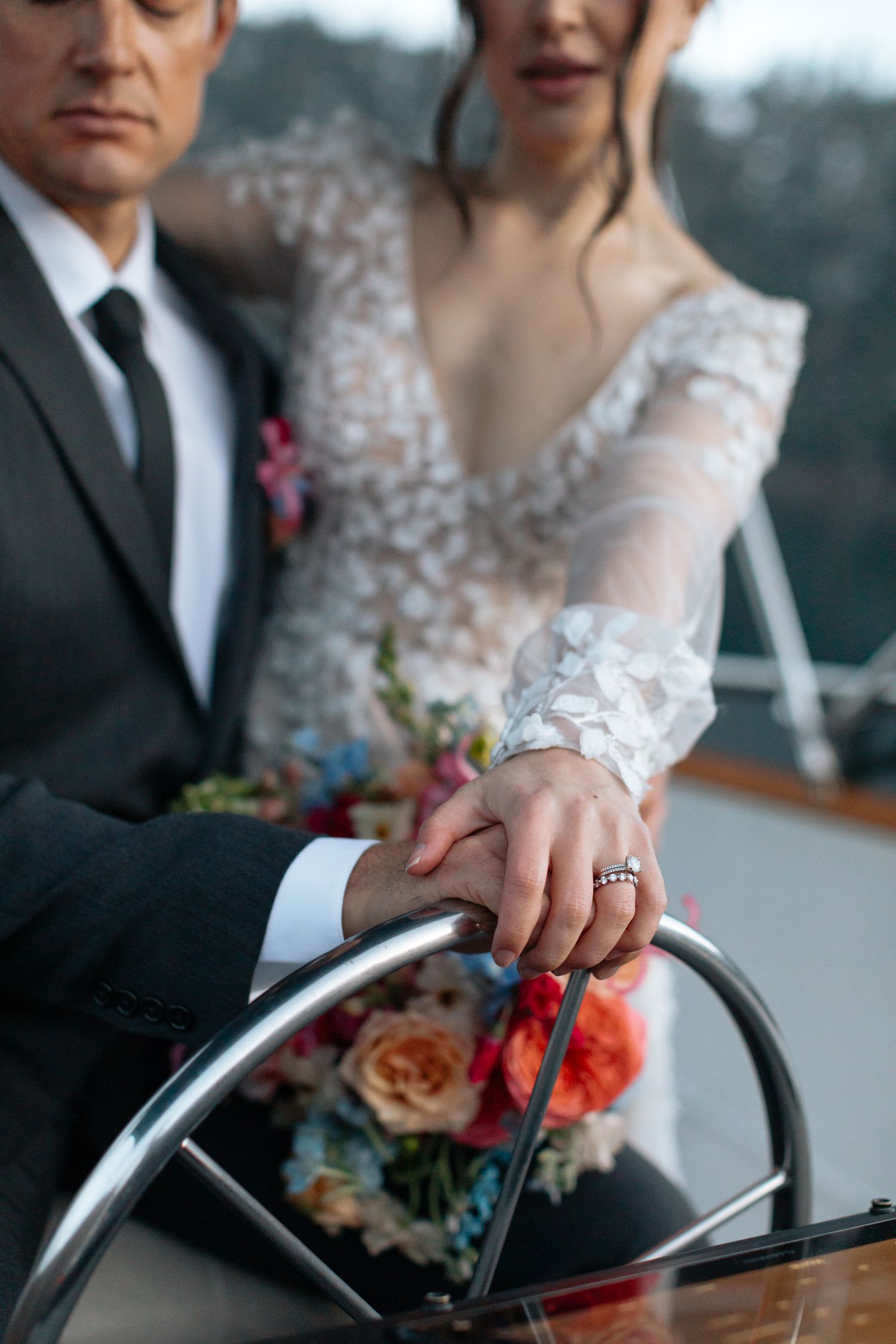 Close-up of hands resting on a boat's steering wheel with wedding rings and floral details.