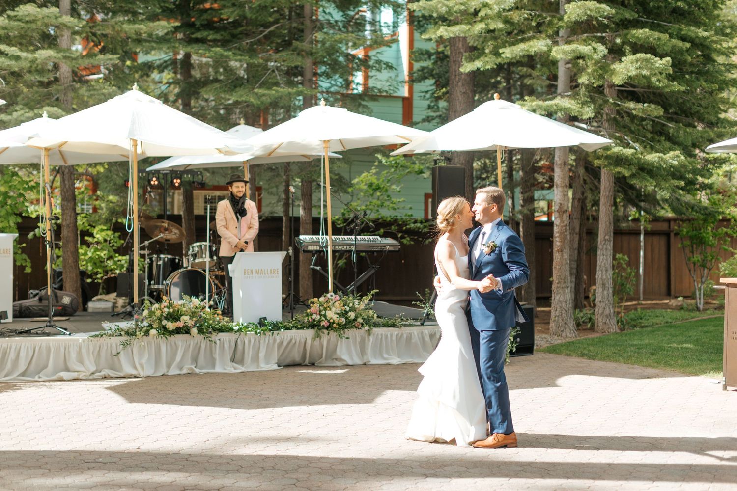 Couples dancing at an outdoor wedding reception under tall pine trees.