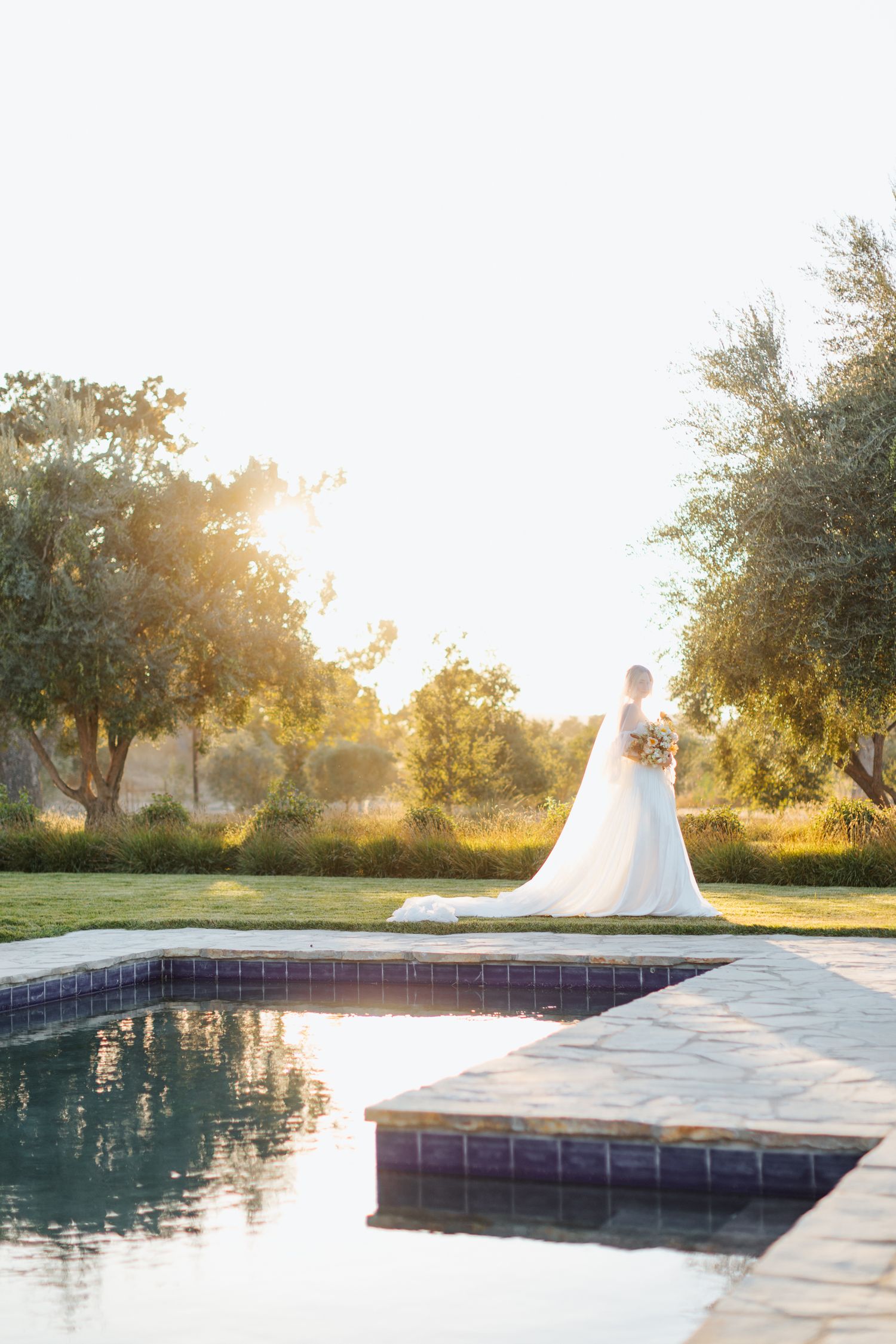 Bride in flowing white dress poses near modern swimming pool during golden hour.