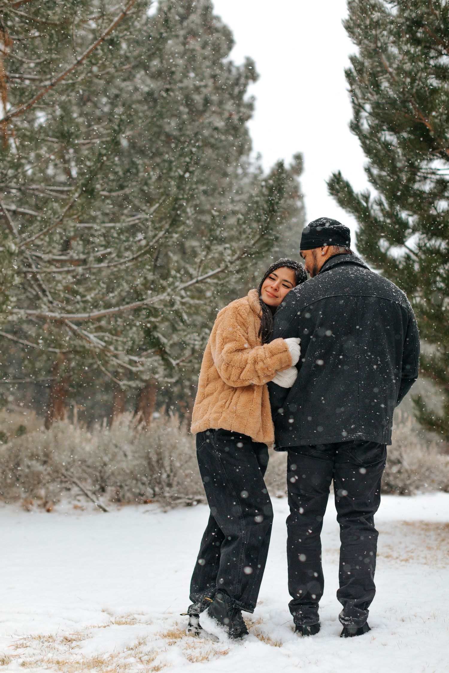 Couple walking together through a snow-covered pine forest in winter clothing.