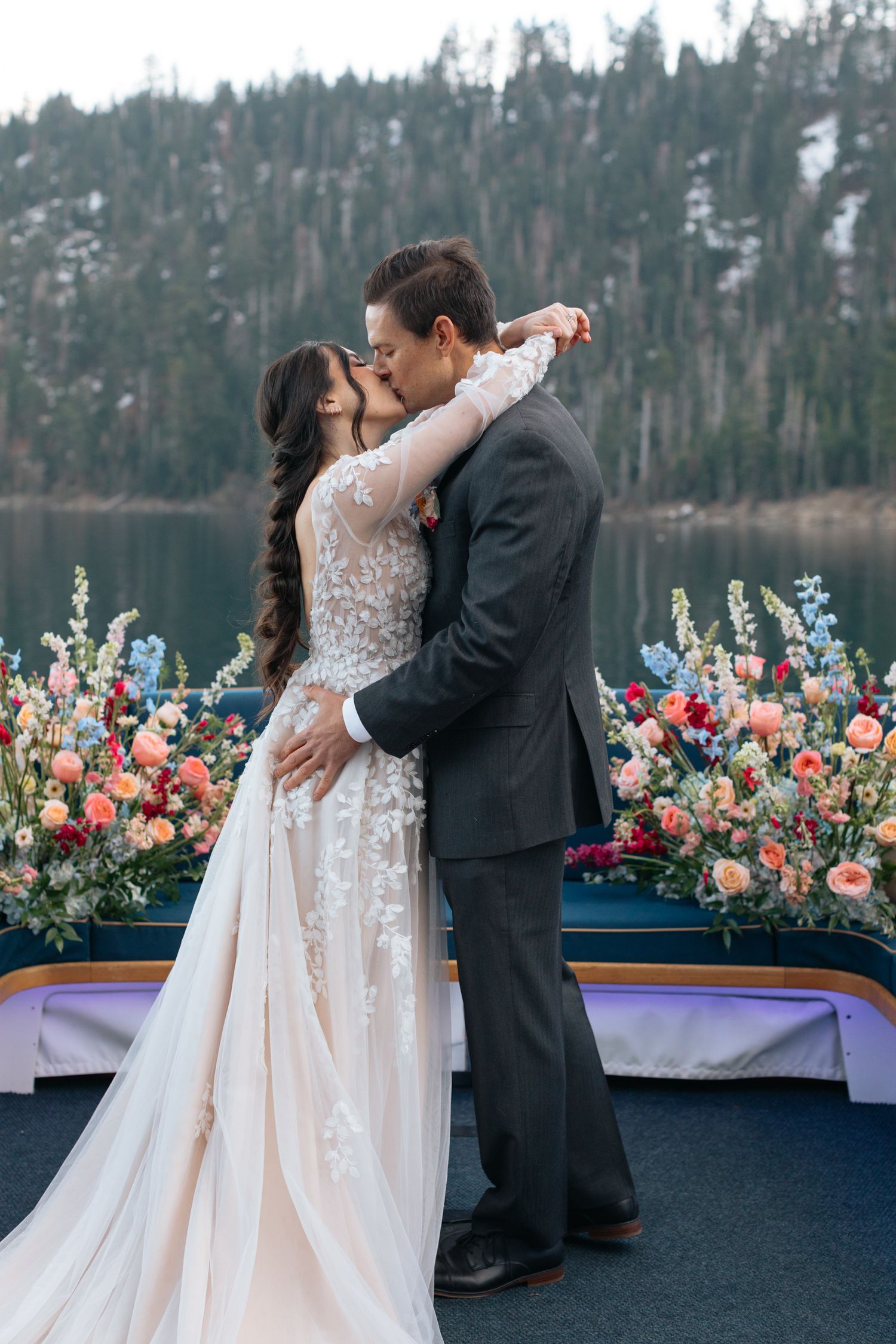Newlyweds dancing on a boat deck with mountain scenery and floral arrangements.
