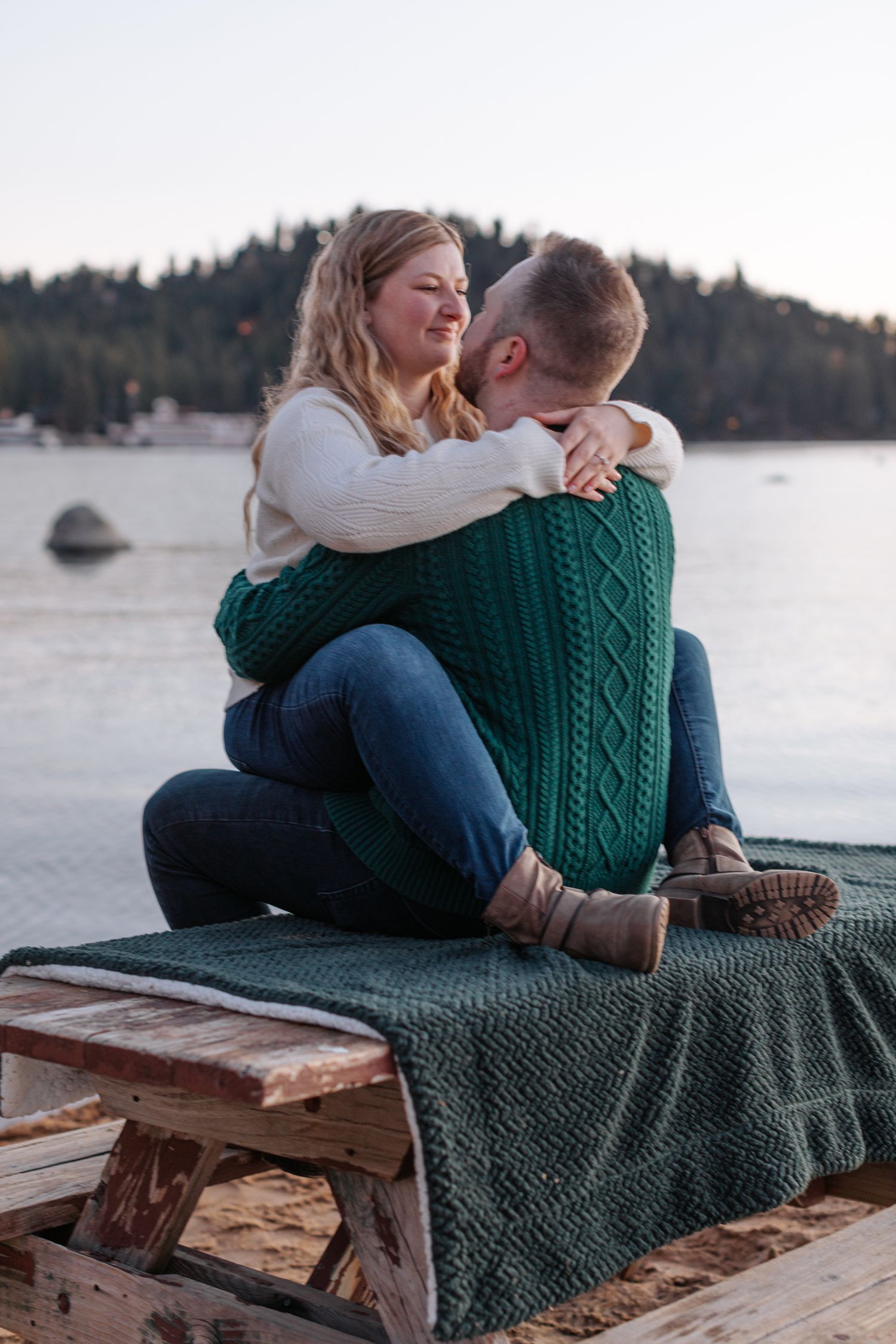 Couple cuddling on wooden bench with green blanket by lakeside during sunset.
