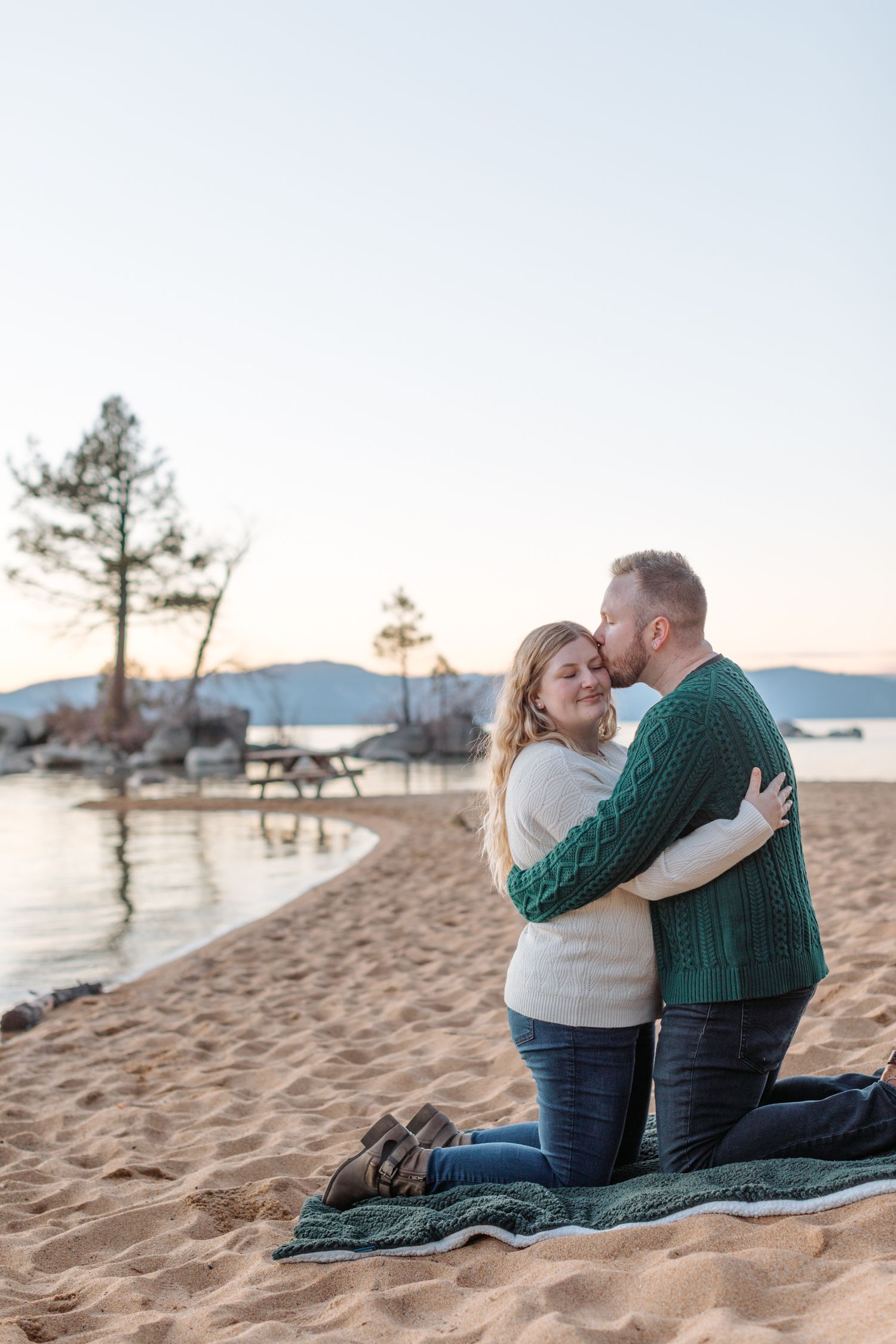 Couple sharing romantic moments on a sandy beach at sunset with mountains and trees in the background.
