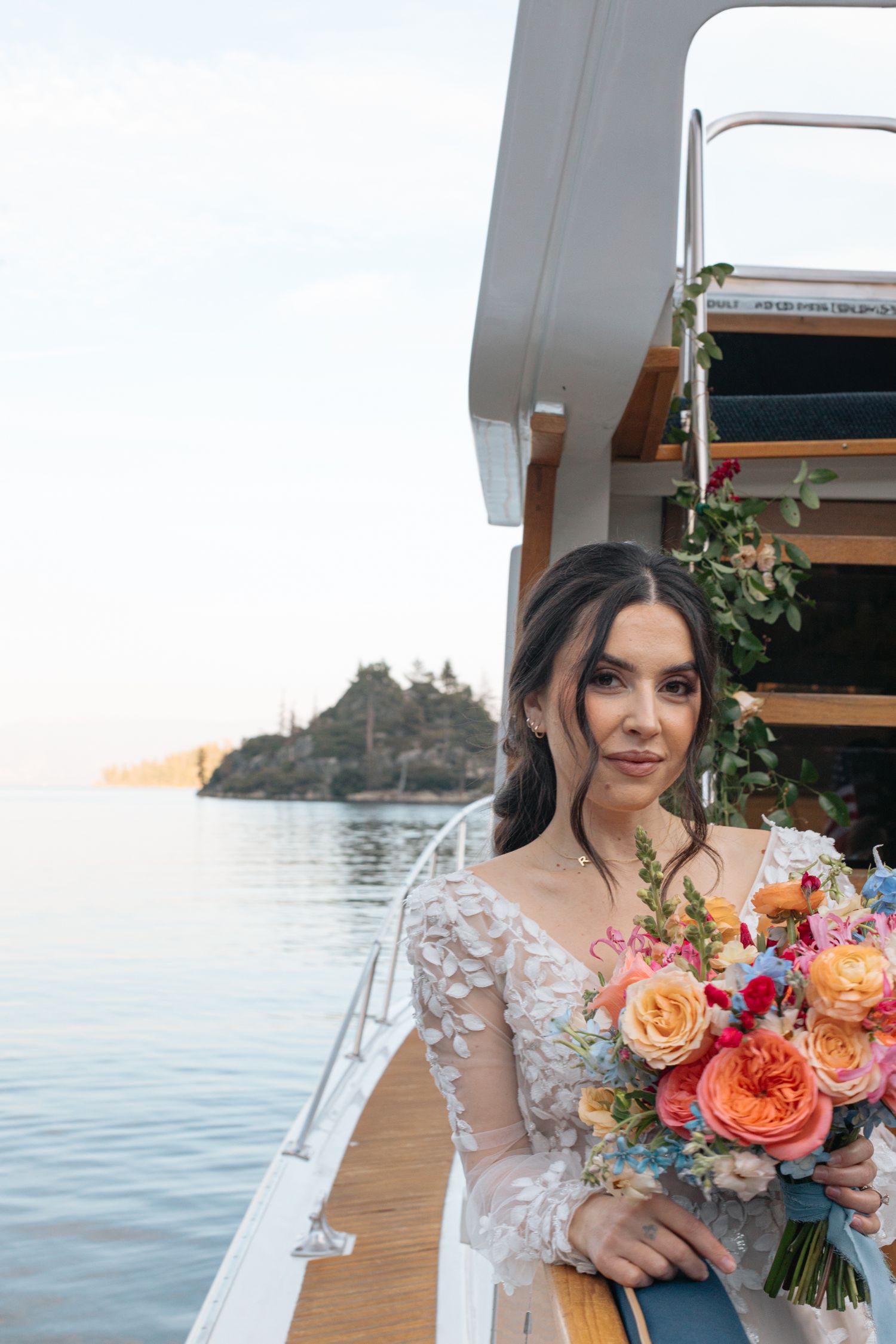 Bride on boat deck holding vibrant bouquet with waterfront and mountains in background.