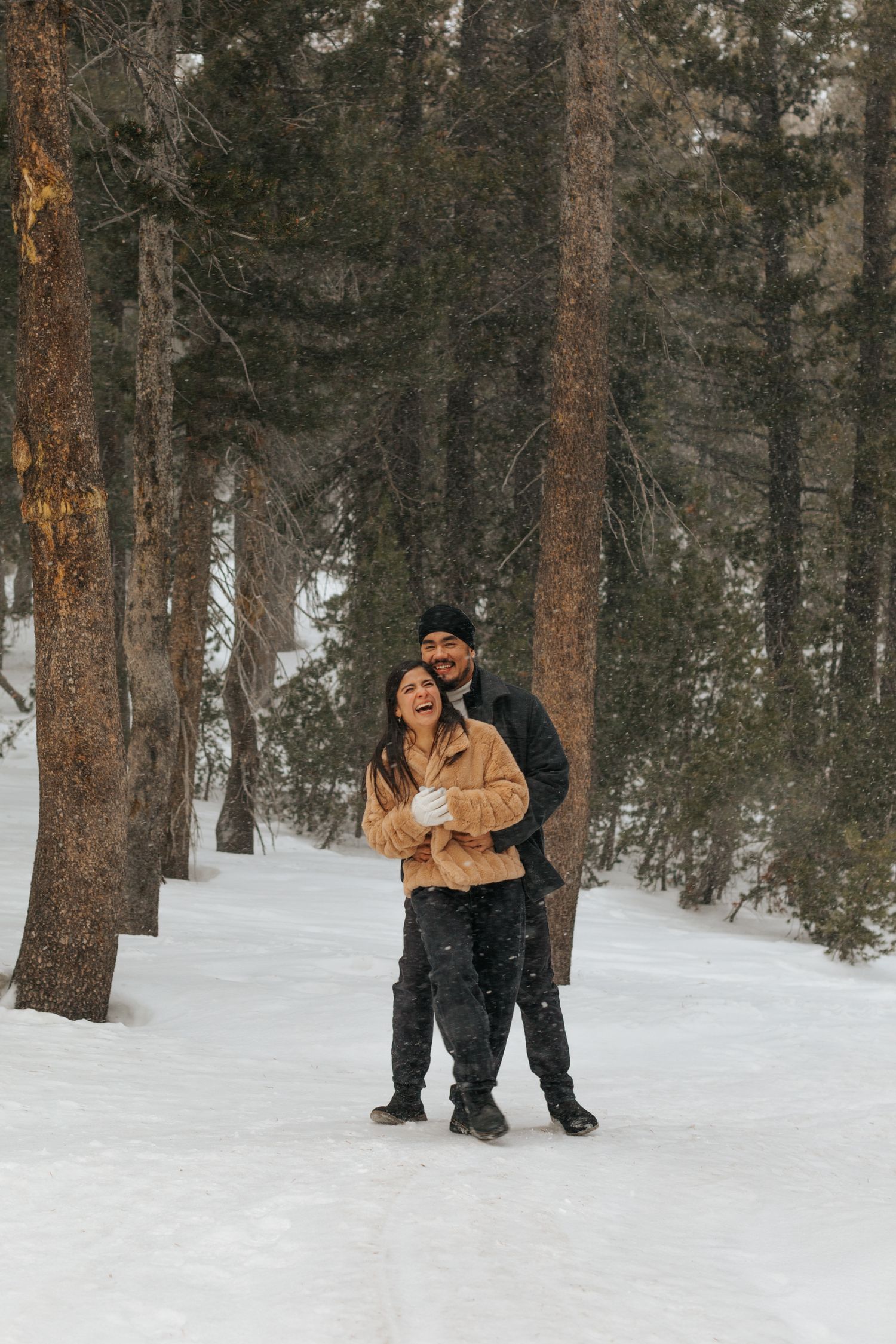 A couple embraces in a snowy forest setting wearing winter coats during a romantic photoshoot.