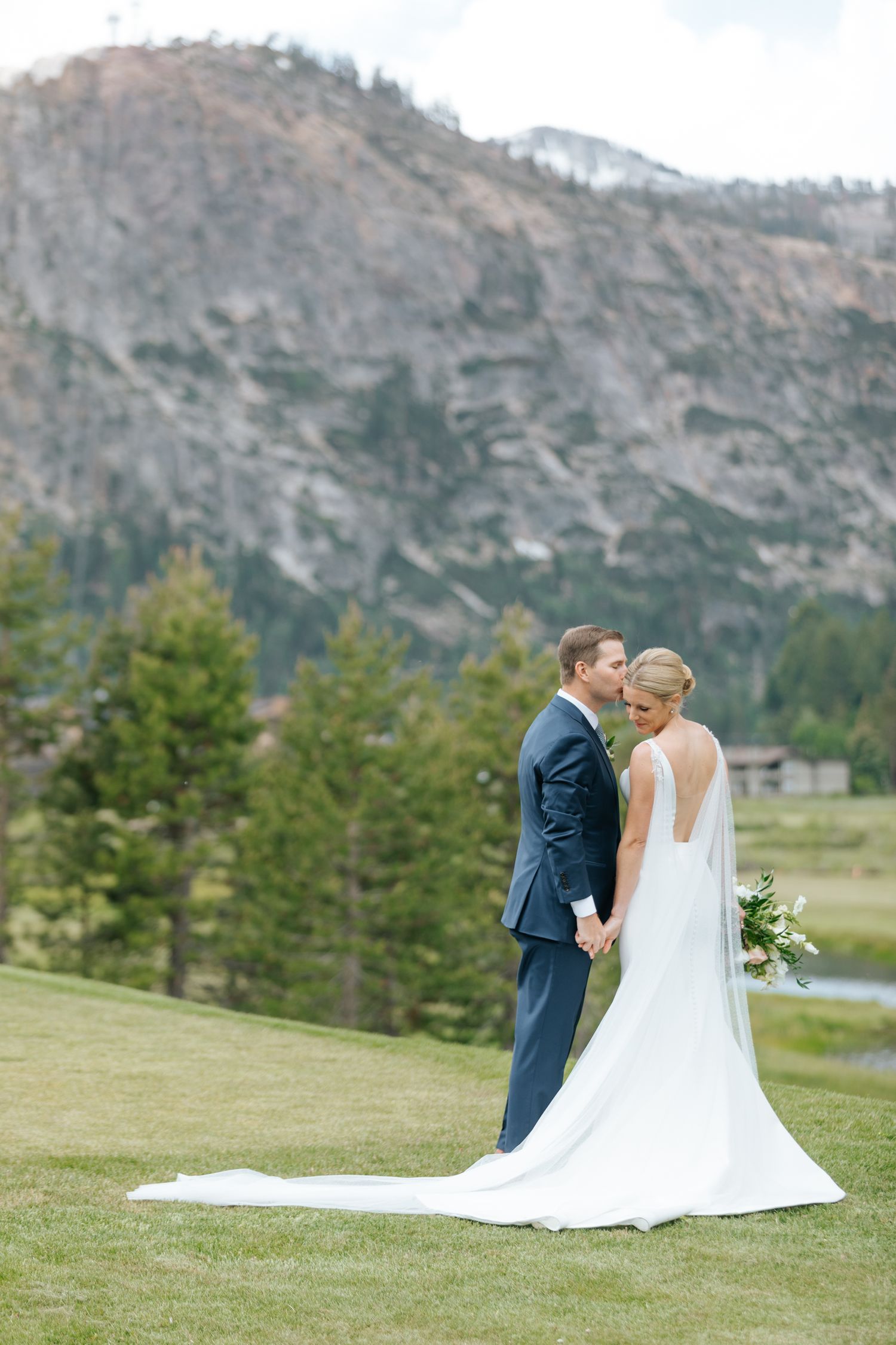 Newlyweds embrace on a grassy hillside with majestic mountain scenery behind them.