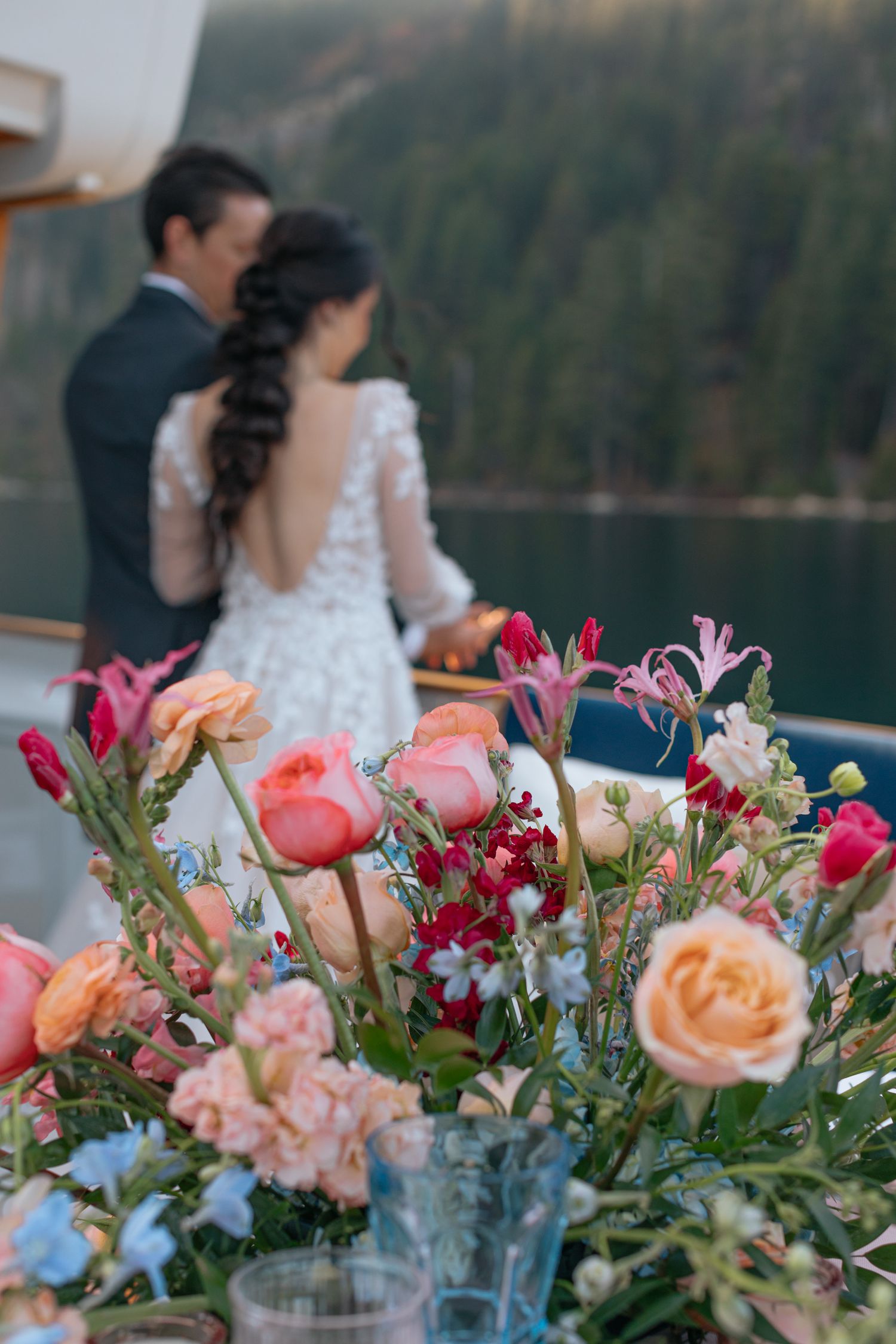 Colorful floral centerpiece with roses and pink flowers while newlyweds stand in background by a lake.