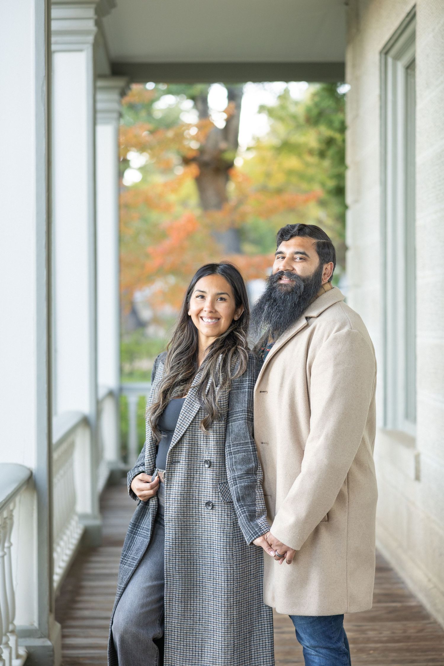 A smiling couple in elegant coats stand on a porch with autumn foliage in the background.