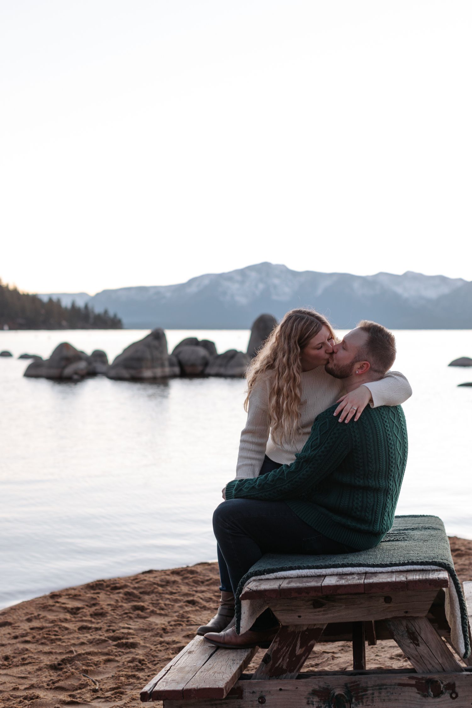 Couple shares intimate moment on wooden bench by mountain lake at sunset.