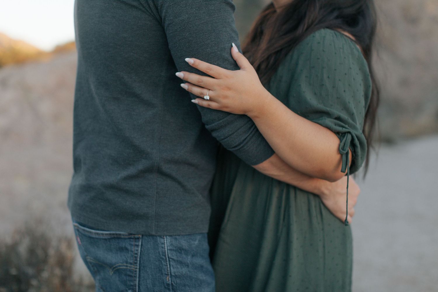 A couple embraces while standing together in a desert landscape with rocky cliffs.