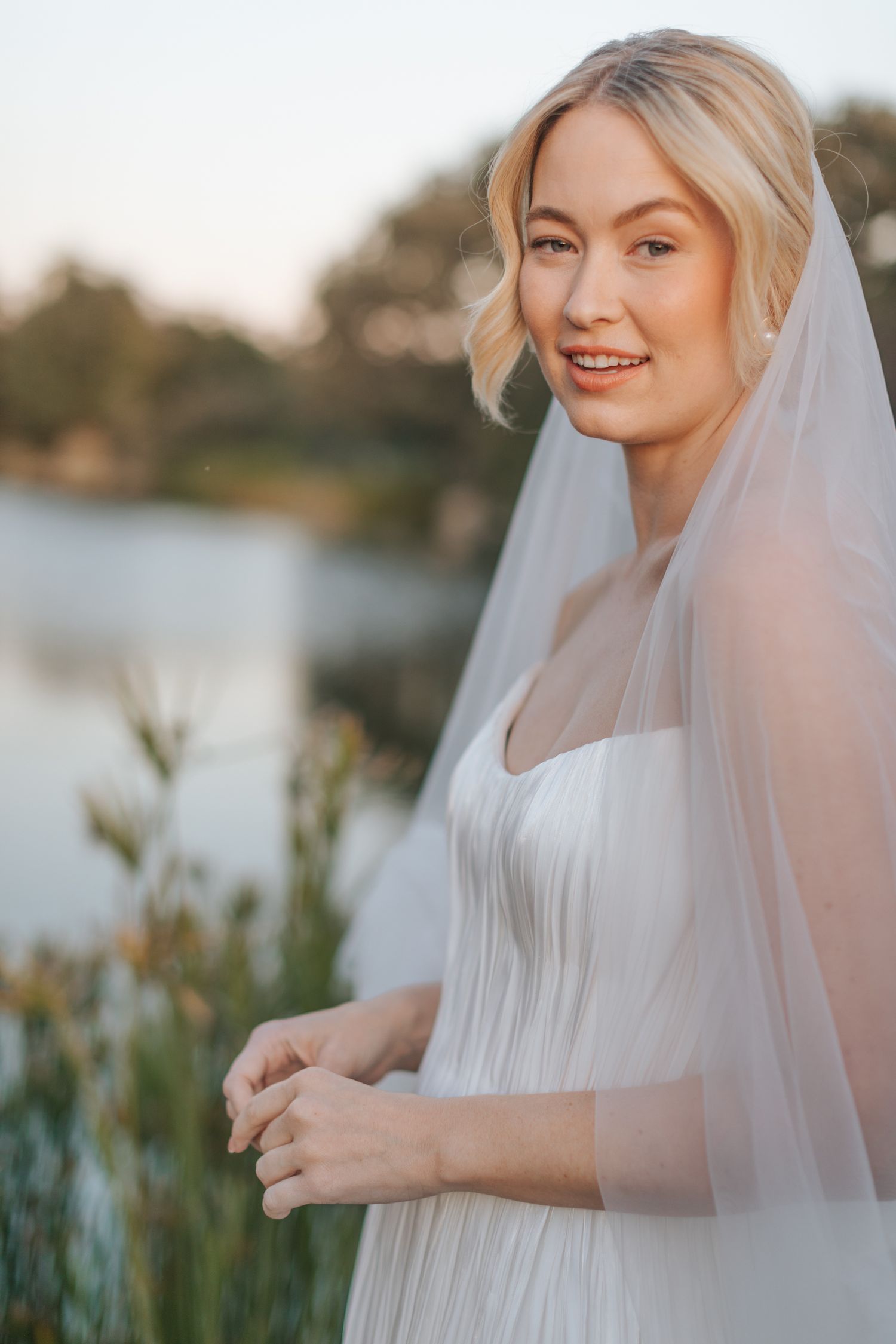 Bride in white dress and veil poses by waterfront during golden hour.