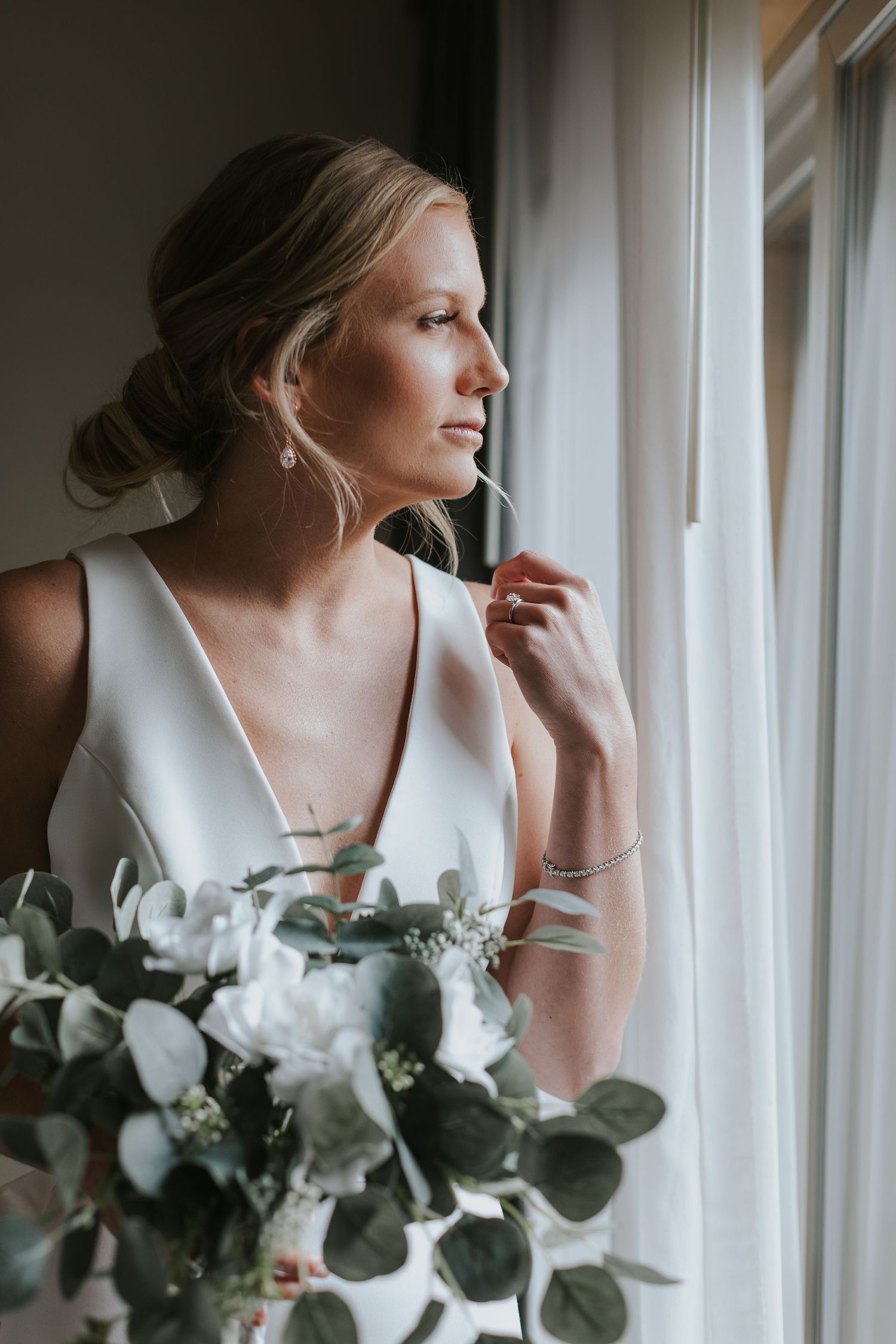 Bride in elegant white dress poses by window with eucalyptus bouquet.