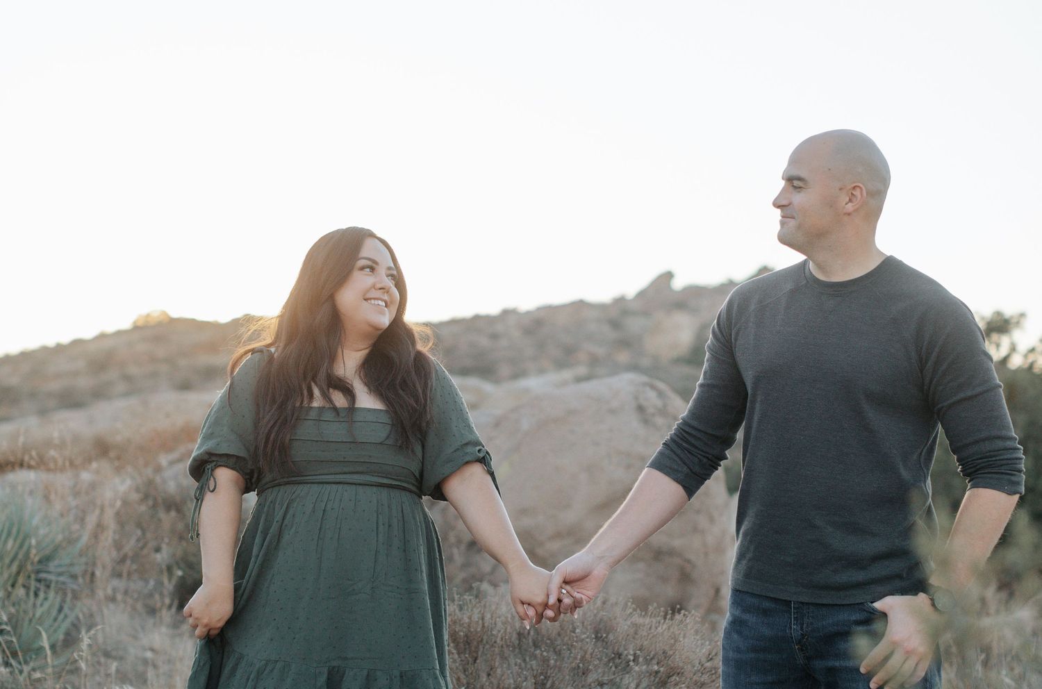 A couple walks hand in hand through a desert landscape at sunset.
