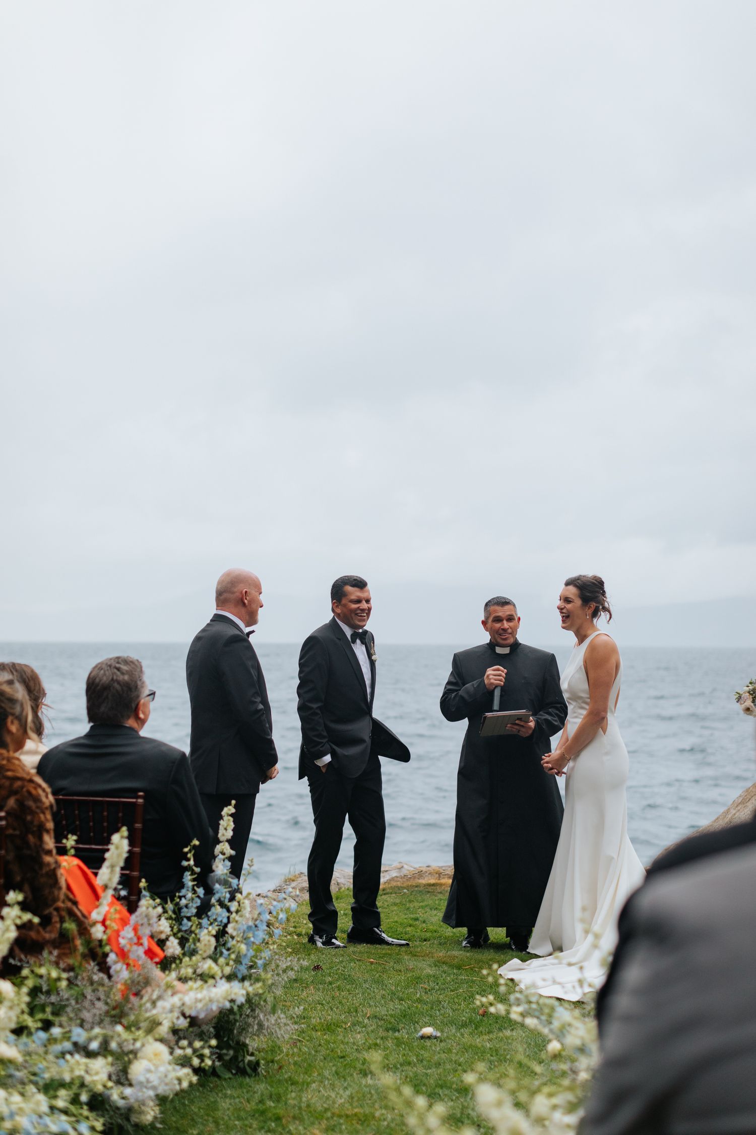 Outdoor wedding ceremony by the ocean with bride, groom, officiant, and guests on a cloudy day.