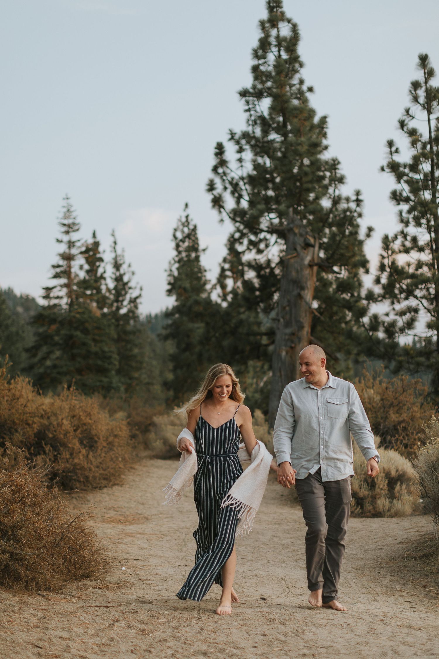 Couple walking hand in hand on a dirt path surrounded by pine trees.