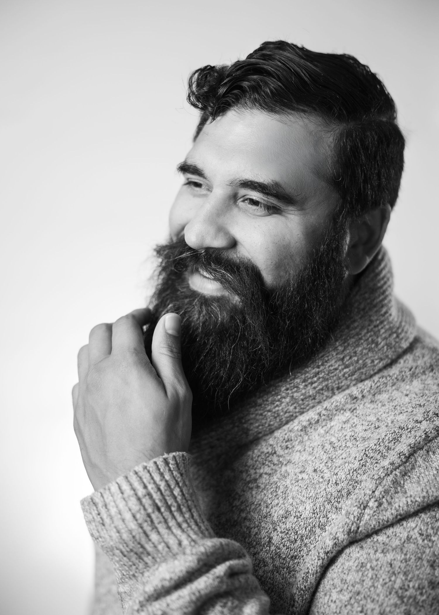 Black and white portrait of a bearded man in a knit sweater looking thoughtfully to the side with hand near chin.