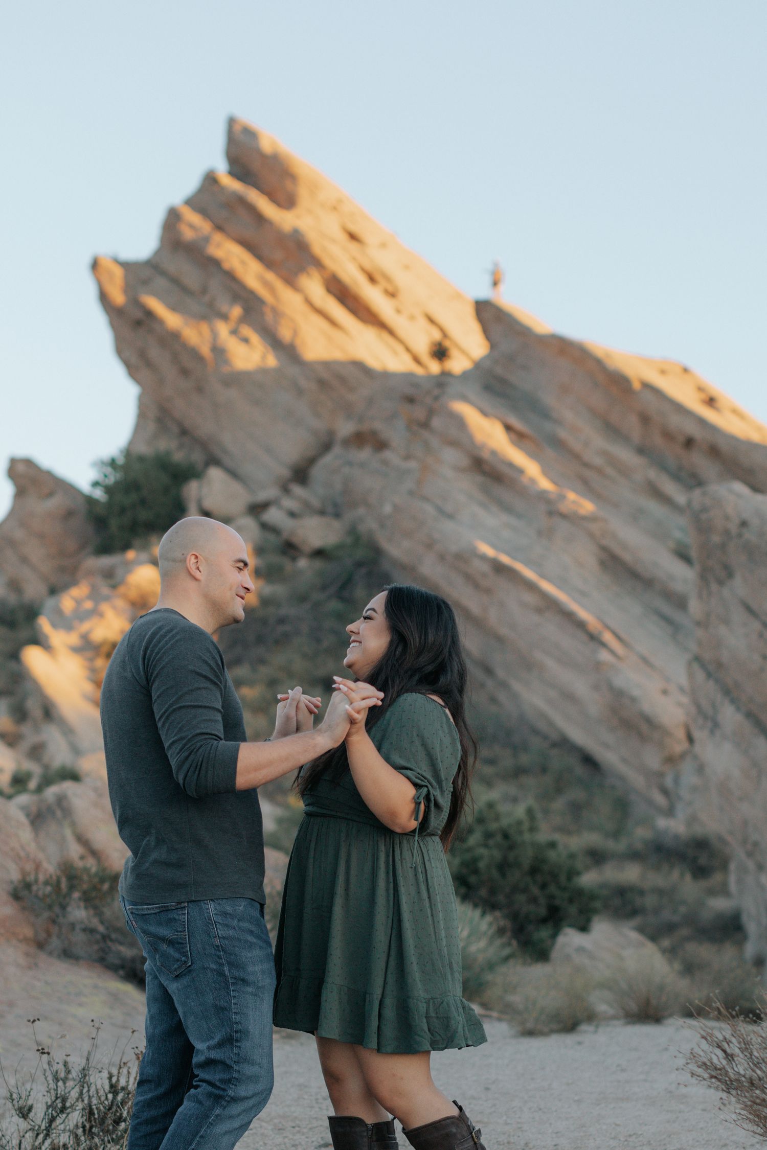 Two people share a romantic moment while standing on a hiking trail near dramatic rock formations.