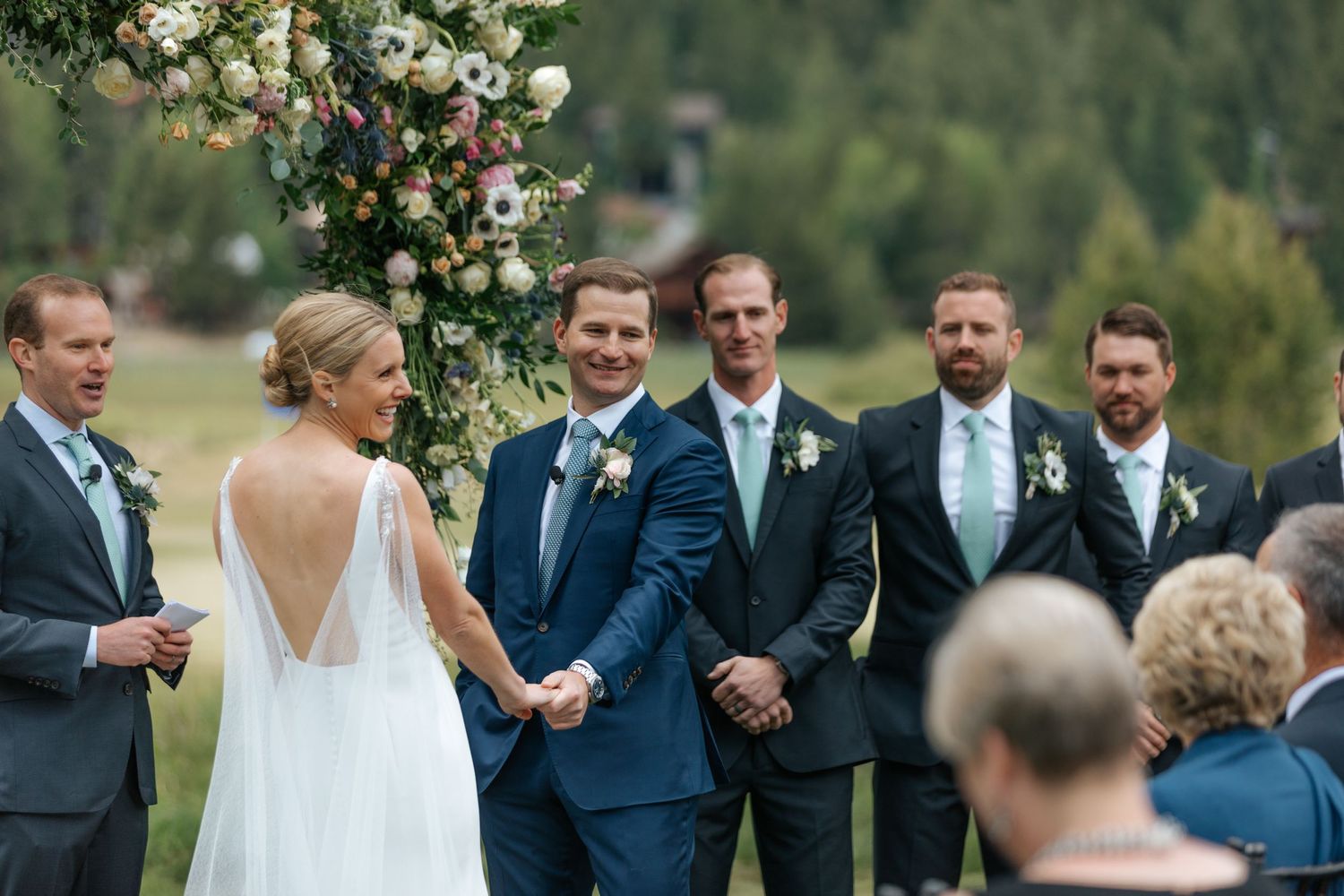 Wedding party stands in formation during outdoor ceremony with floral arch and greenery.