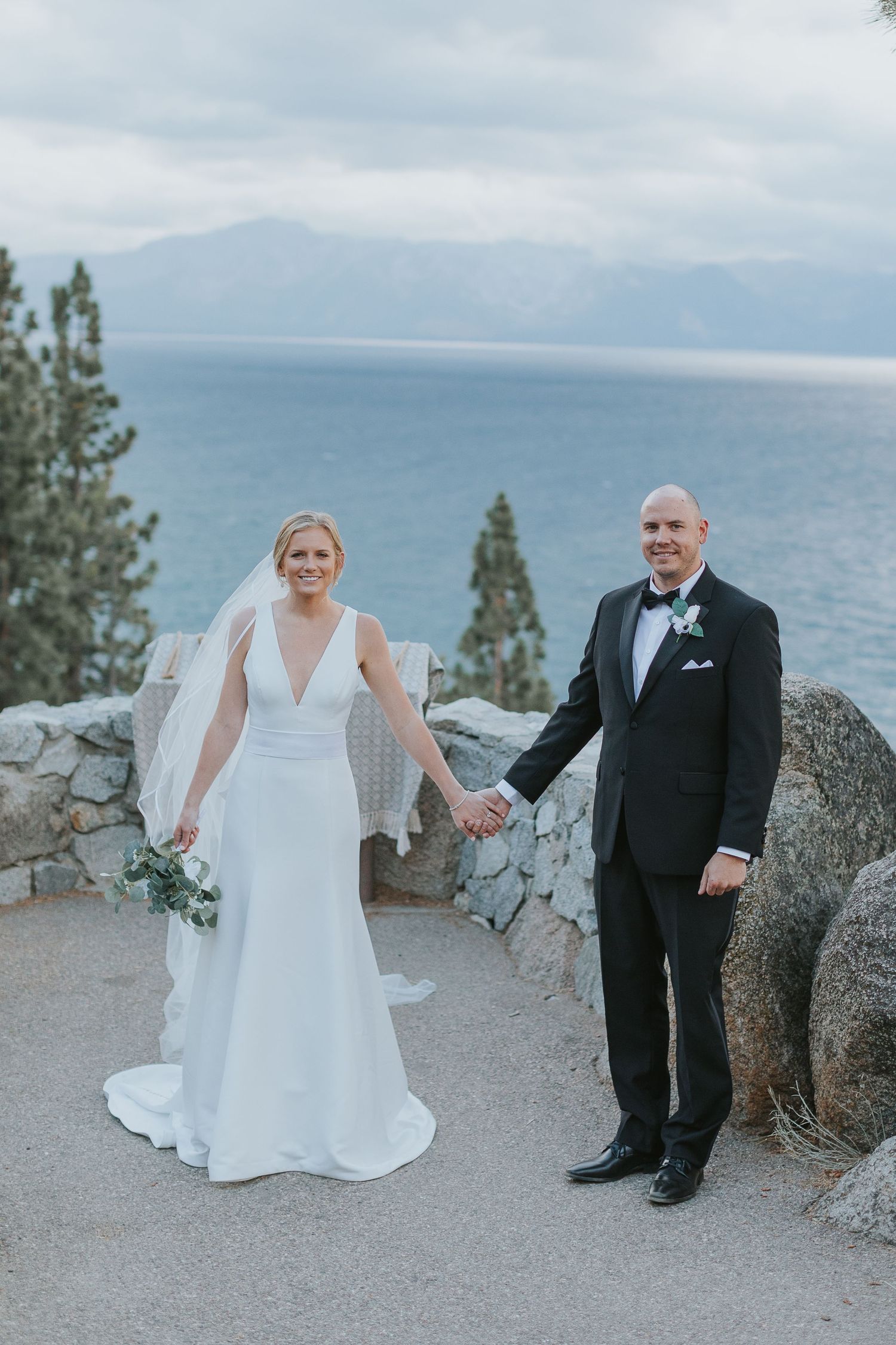 A wedding couple poses against a scenic mountain backdrop overlooking a lake.