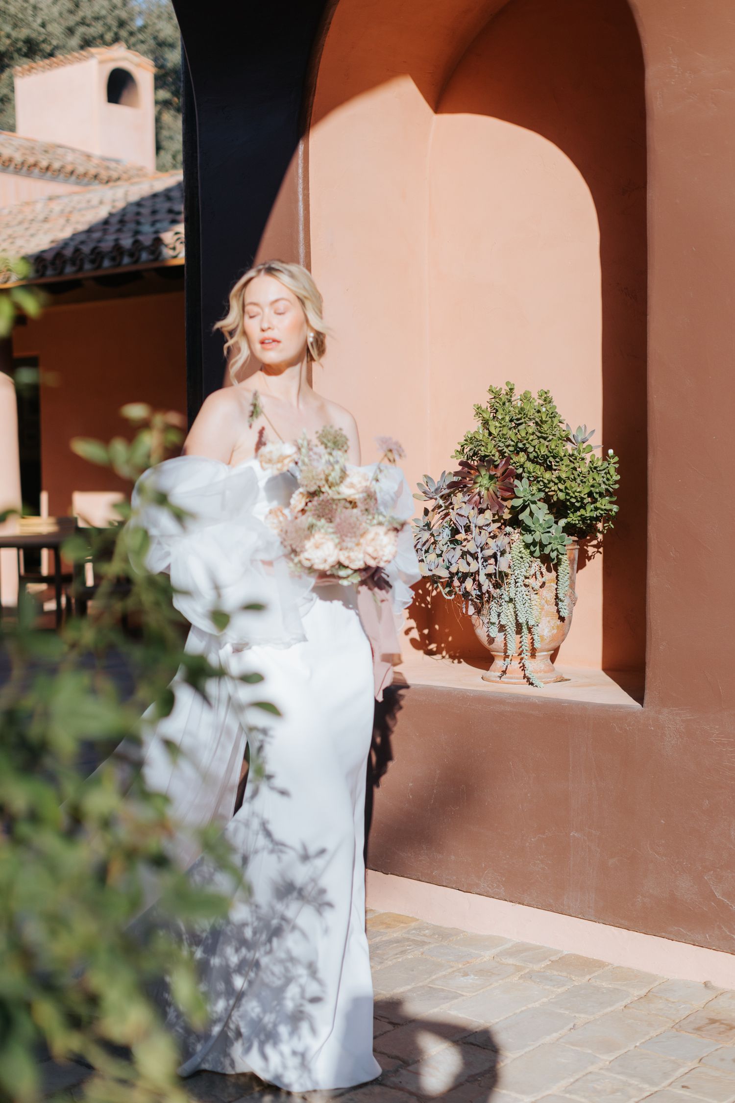 A bride in a white dress poses against a peach-colored archway surrounded by potted plants.