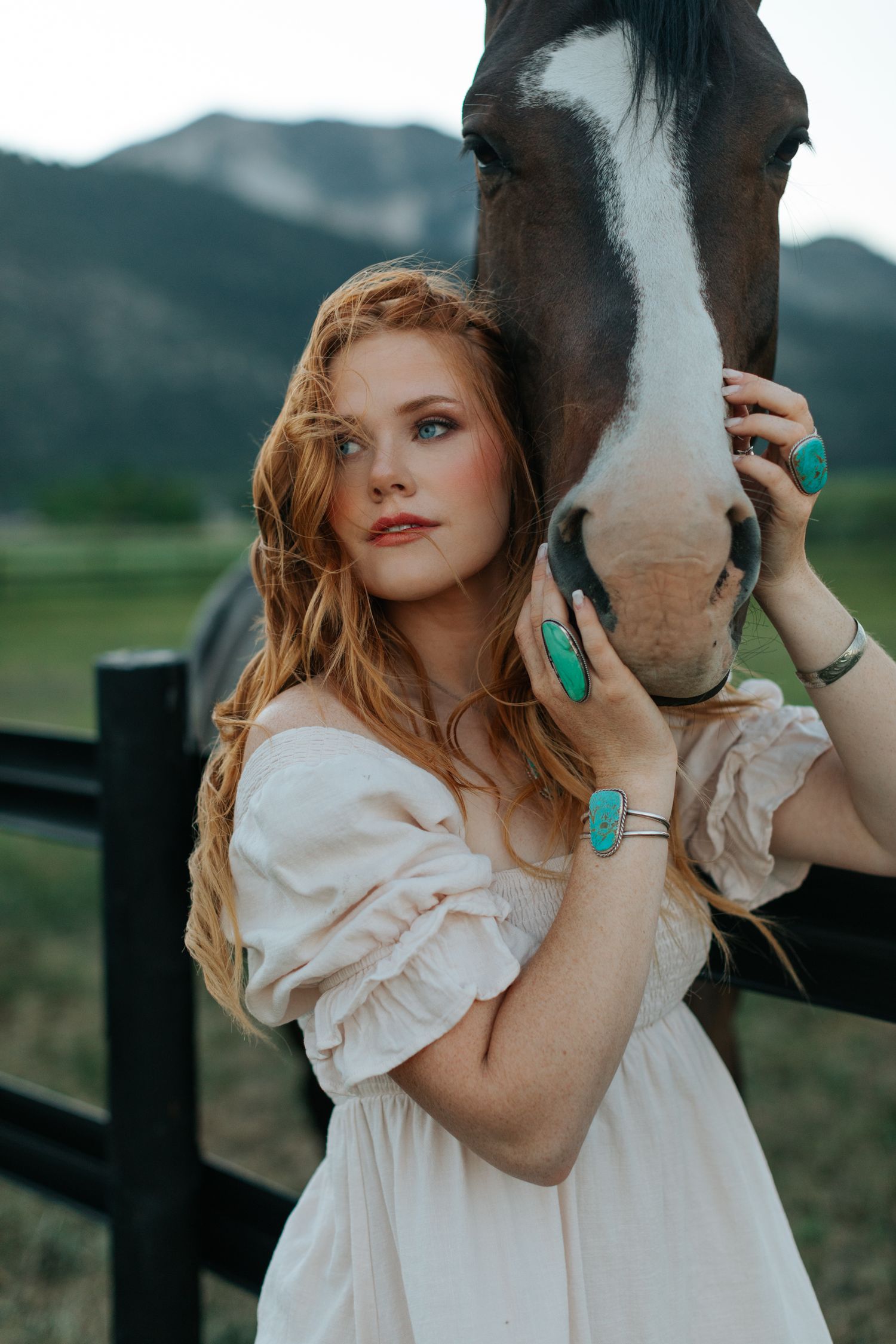 A woman in a white dress embraces a horse's face against a blurred rural landscape background.
