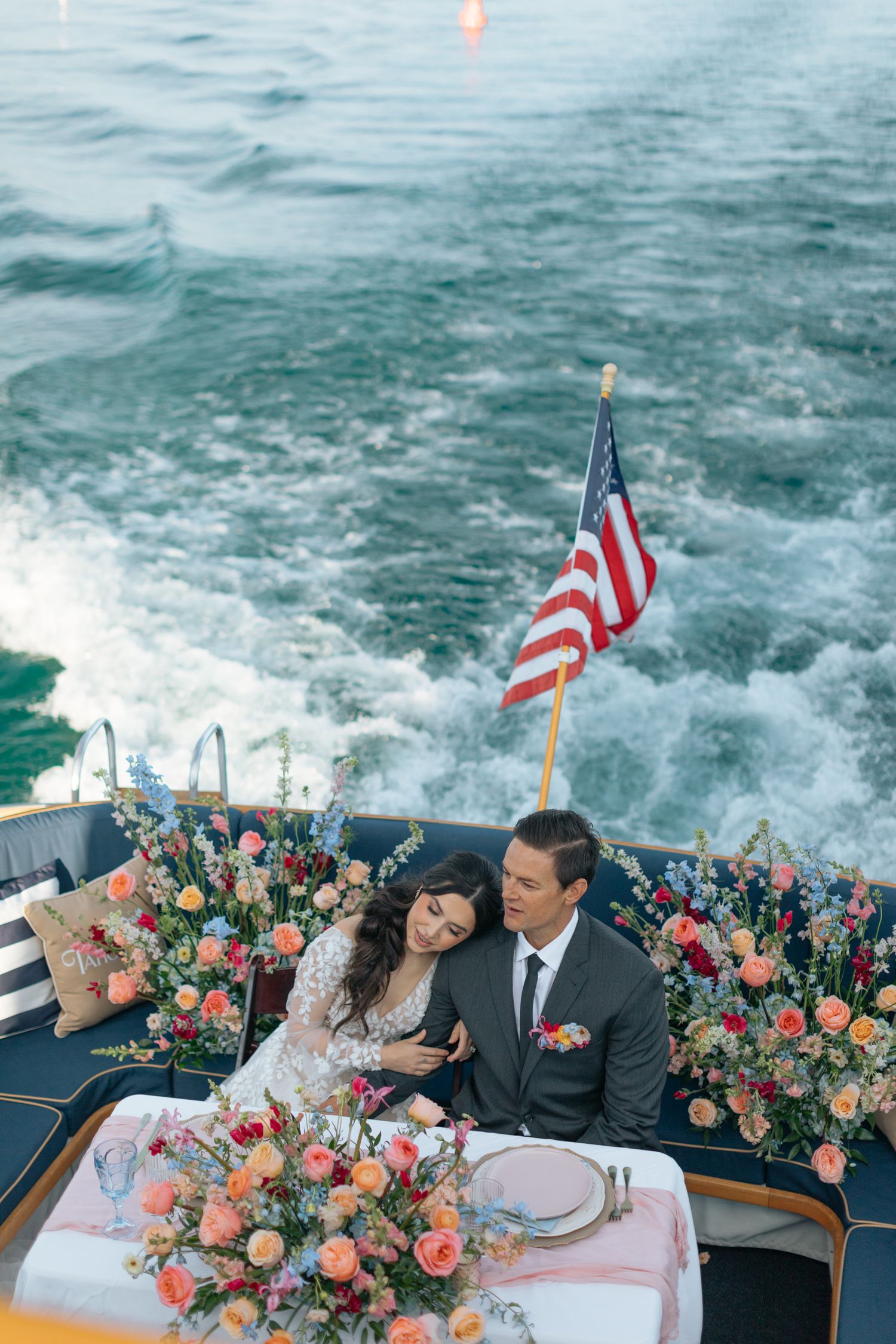 Couple in wedding attire seated at floral table on boat stern with American flag and ocean wake backdrop.