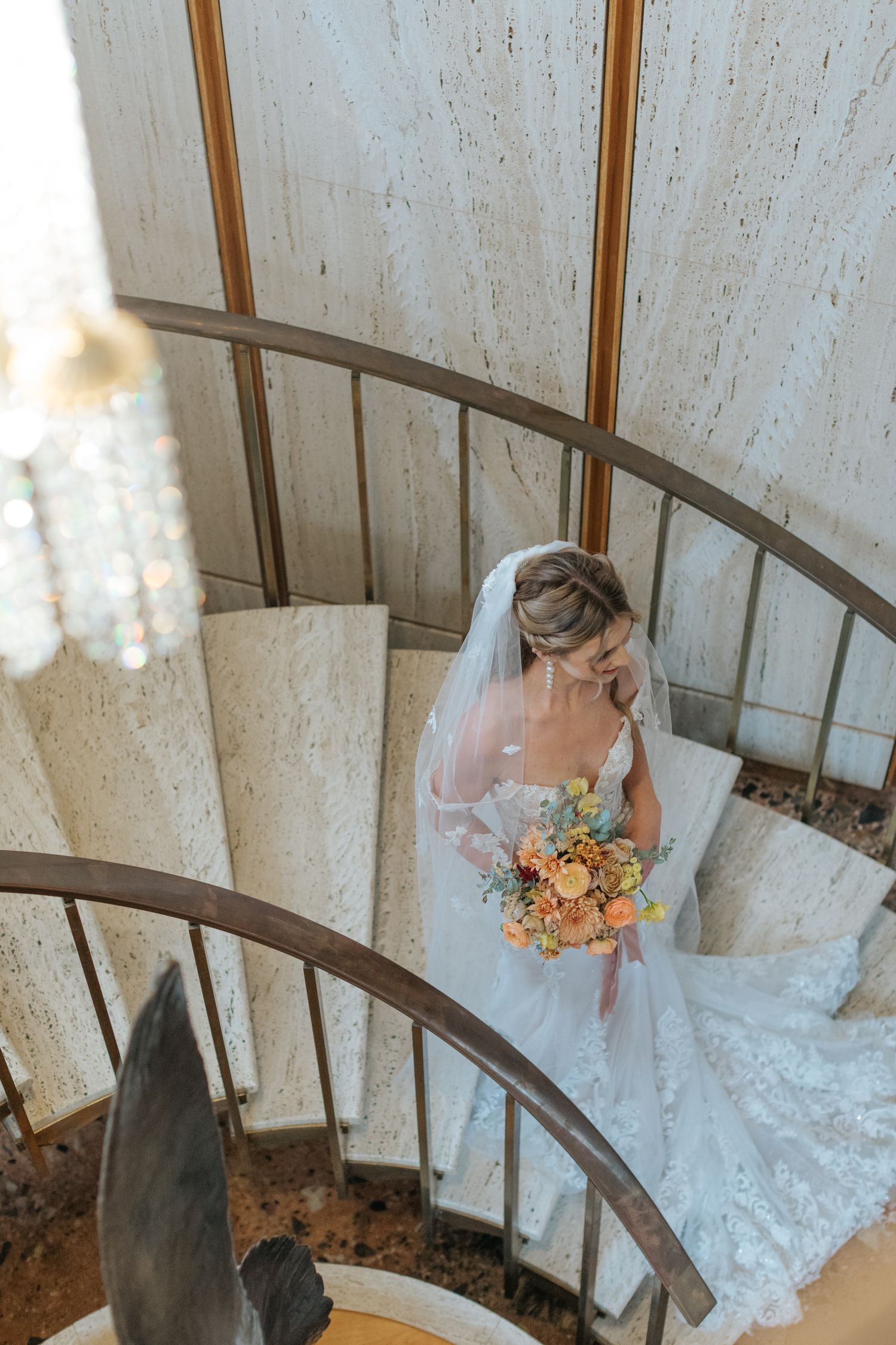 White lace wedding dress flowing down curved wooden staircase with autumn-colored bouquet.