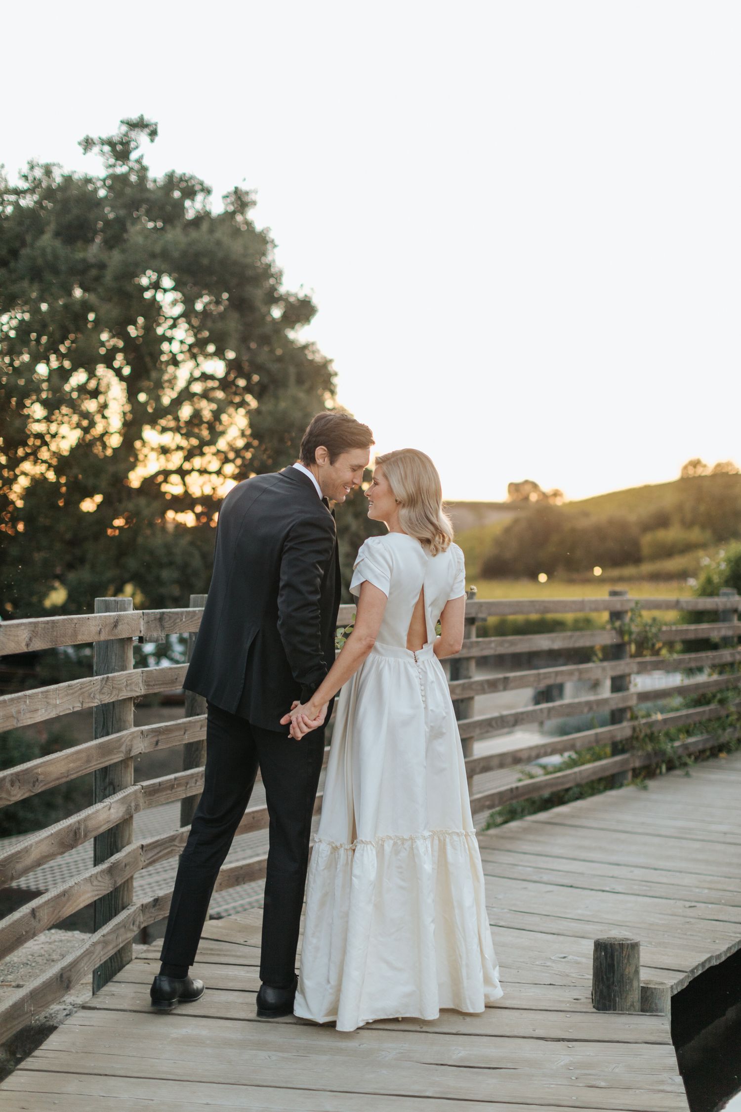 A couple in wedding attire shares a romantic moment on a wooden bridge at sunset.