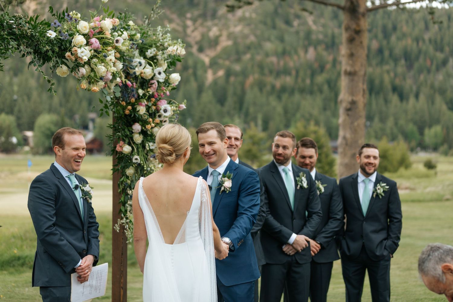 Wedding party in navy suits and white dress gather for outdoor ceremony against mountain backdrop.