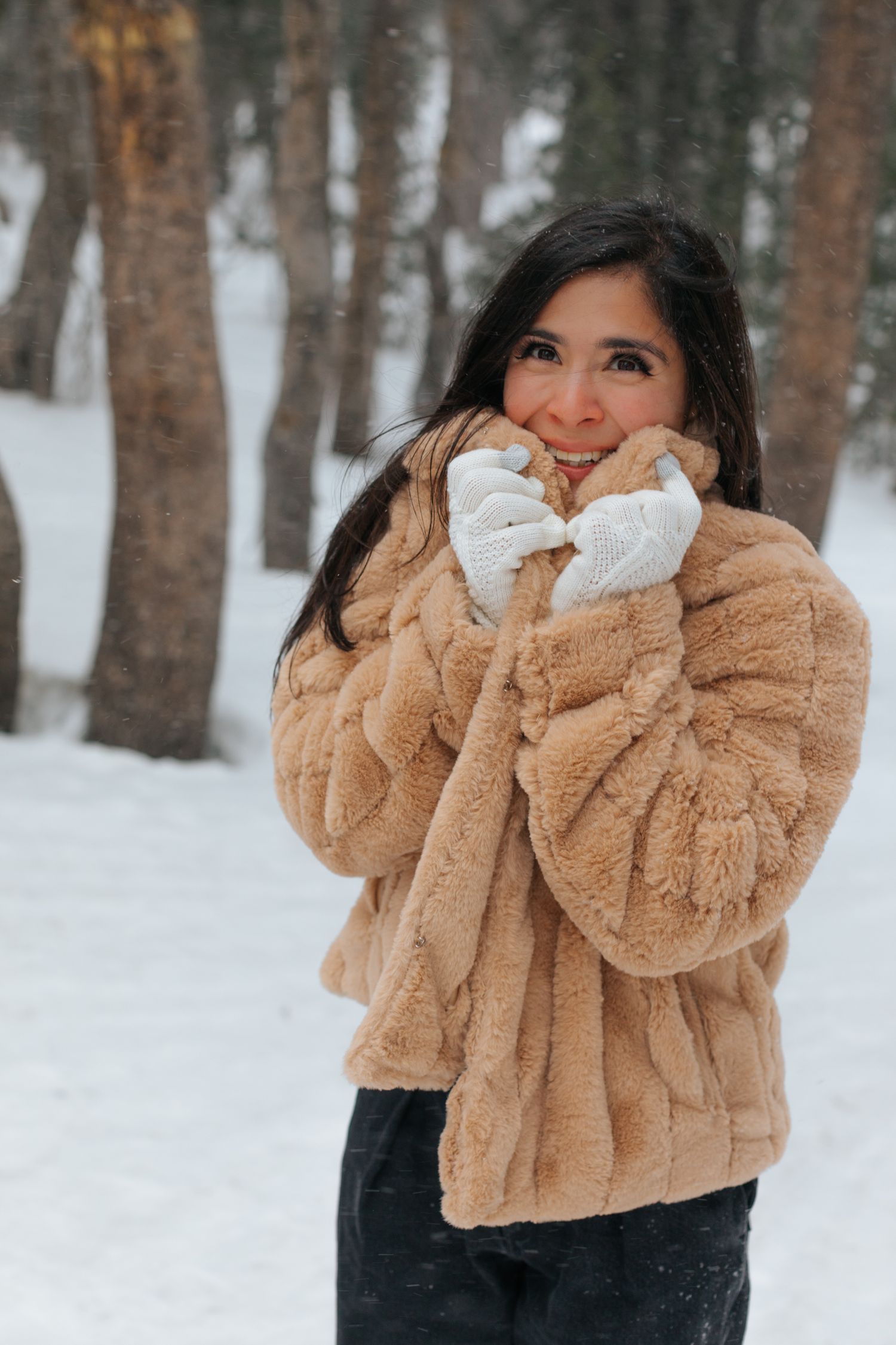 Person in a cozy beige faux fur coat poses playfully in a snowy winter forest.