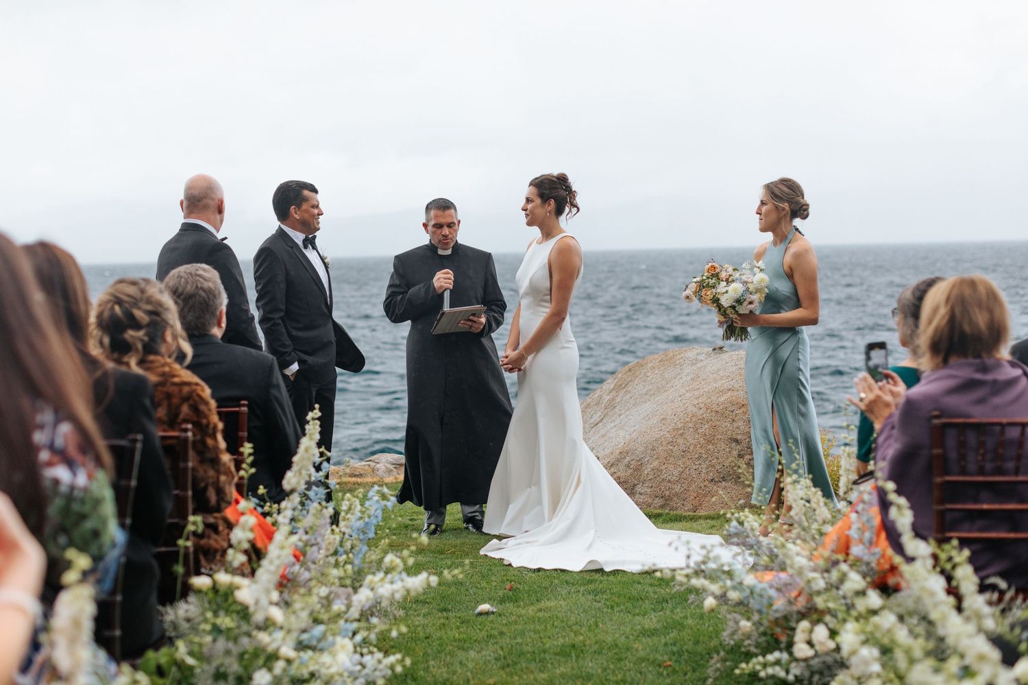 Oceanside wedding ceremony setup with floral arrangements and guests gathering by the water.