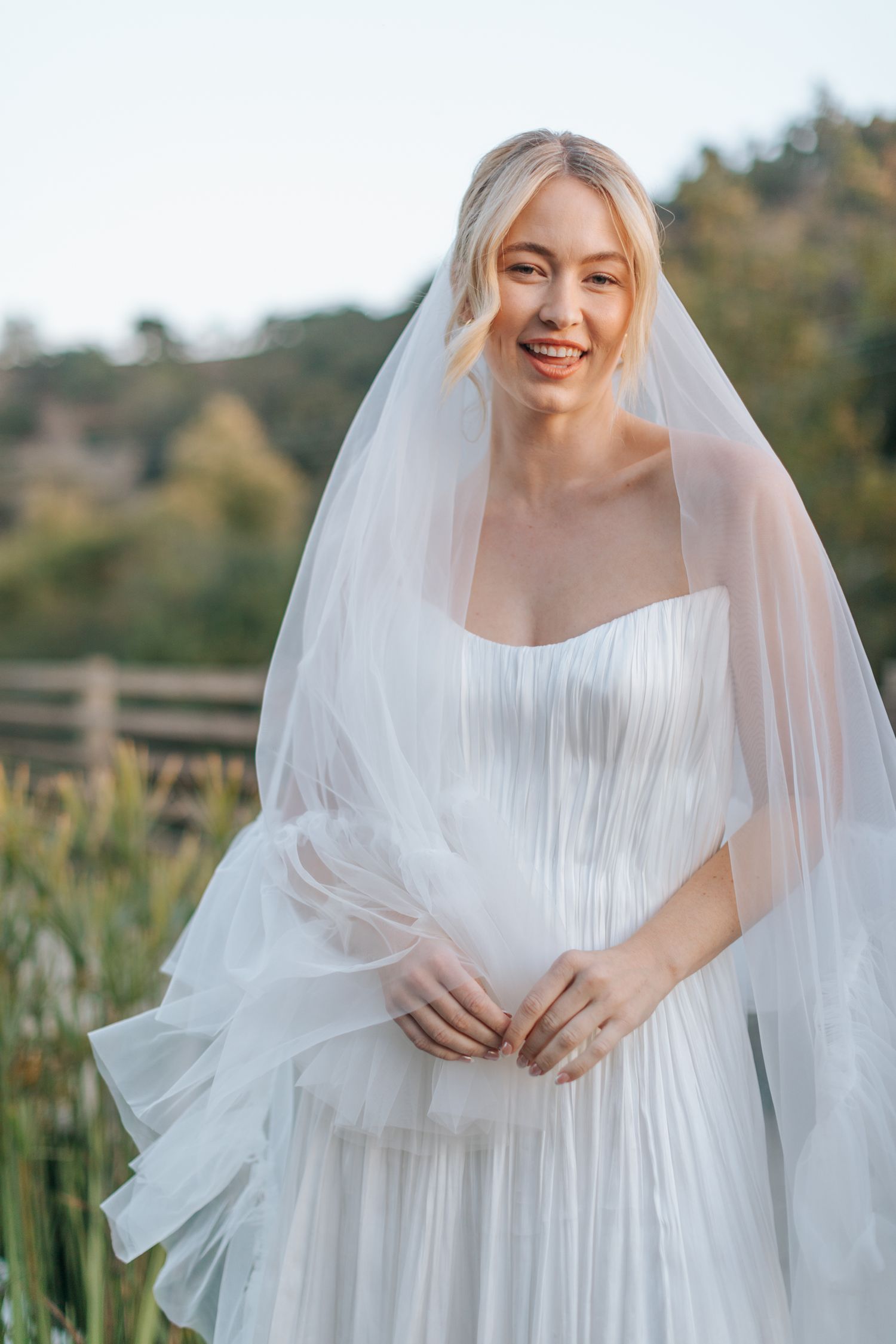 A bride in a flowing white pleated dress and veil poses outdoors in a natural setting during a wedding photoshoot.
