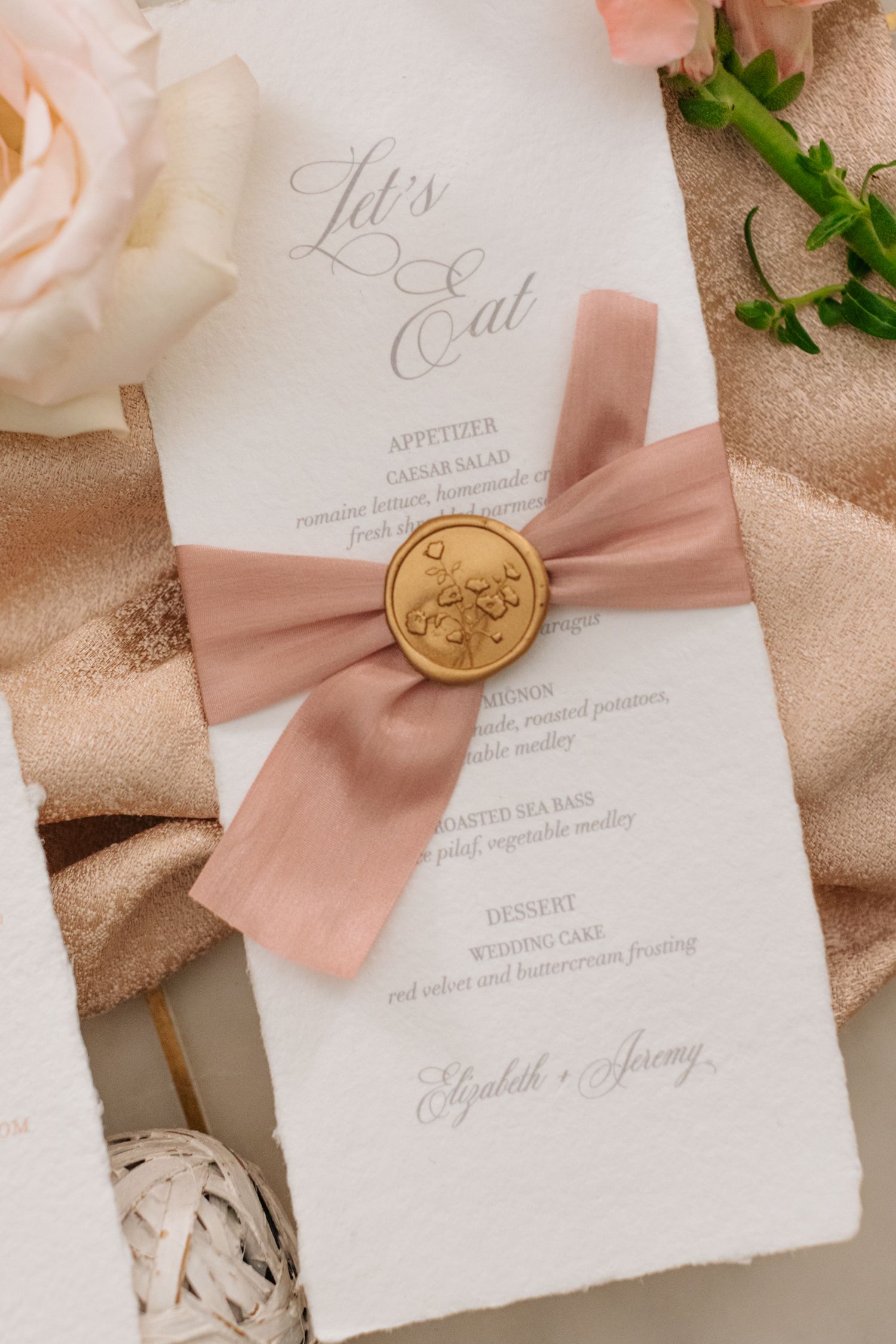 Wedding menu cards with pink silk ribbons and gold wax seals arranged with roses.
