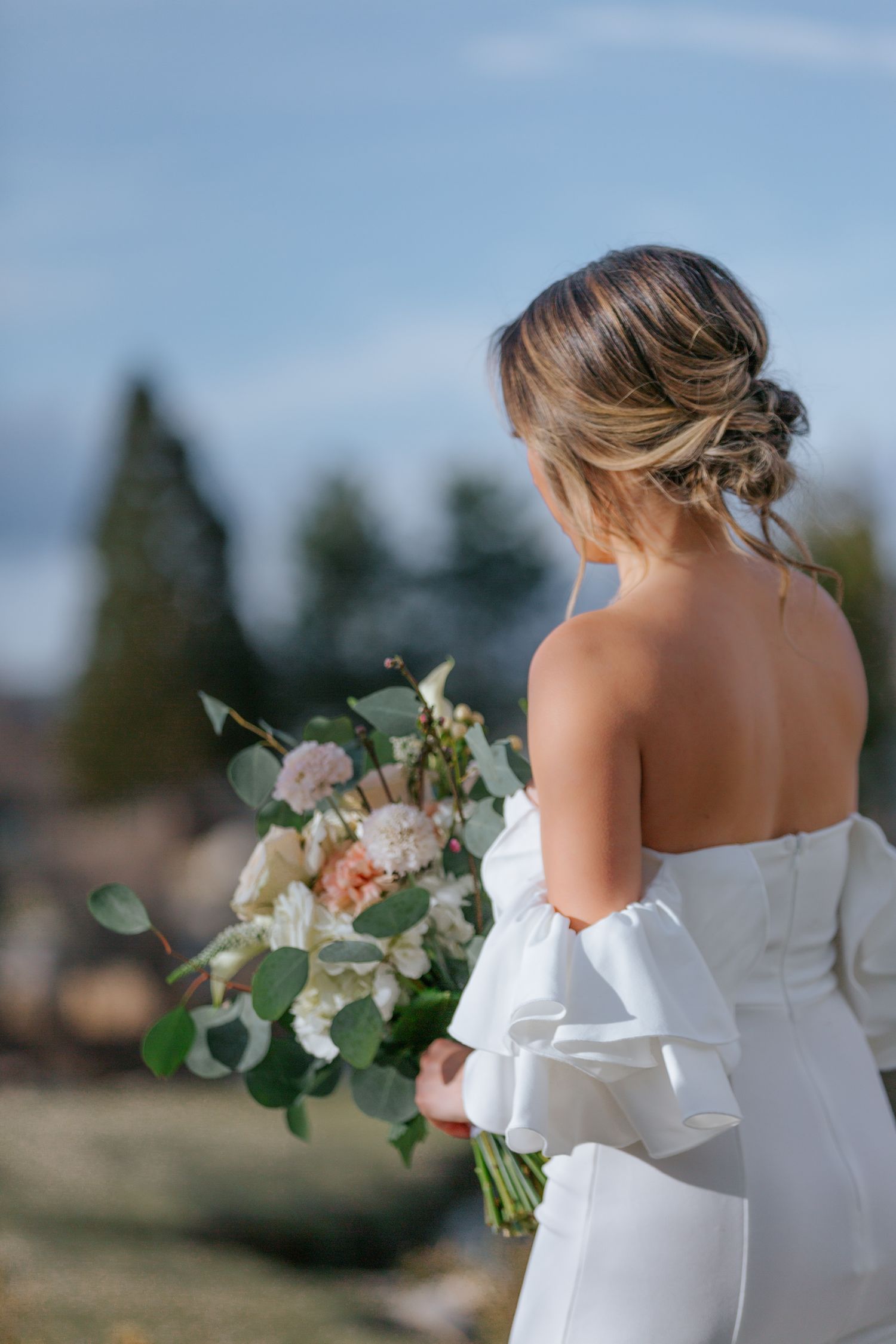An elegant white wedding dress with floral bouquet against a natural outdoor backdrop.