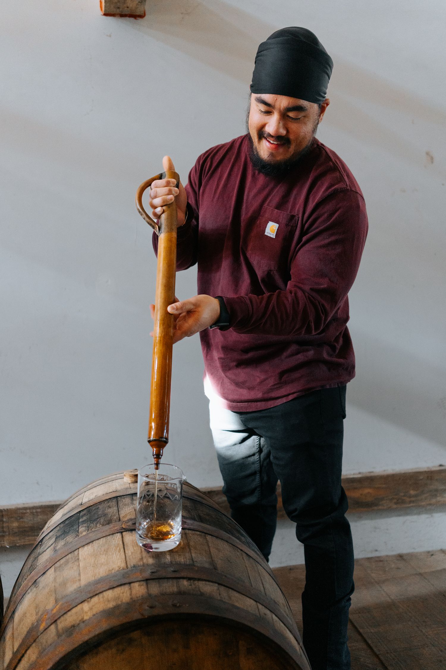 Worker extracting bourbon samples from barrel using a whiskey thief tool.