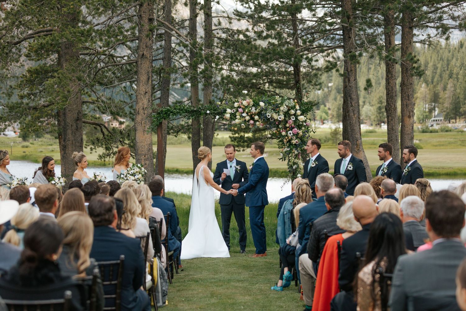 Scenic outdoor wedding ceremony surrounded by tall pine trees with guests seated on white chairs.
