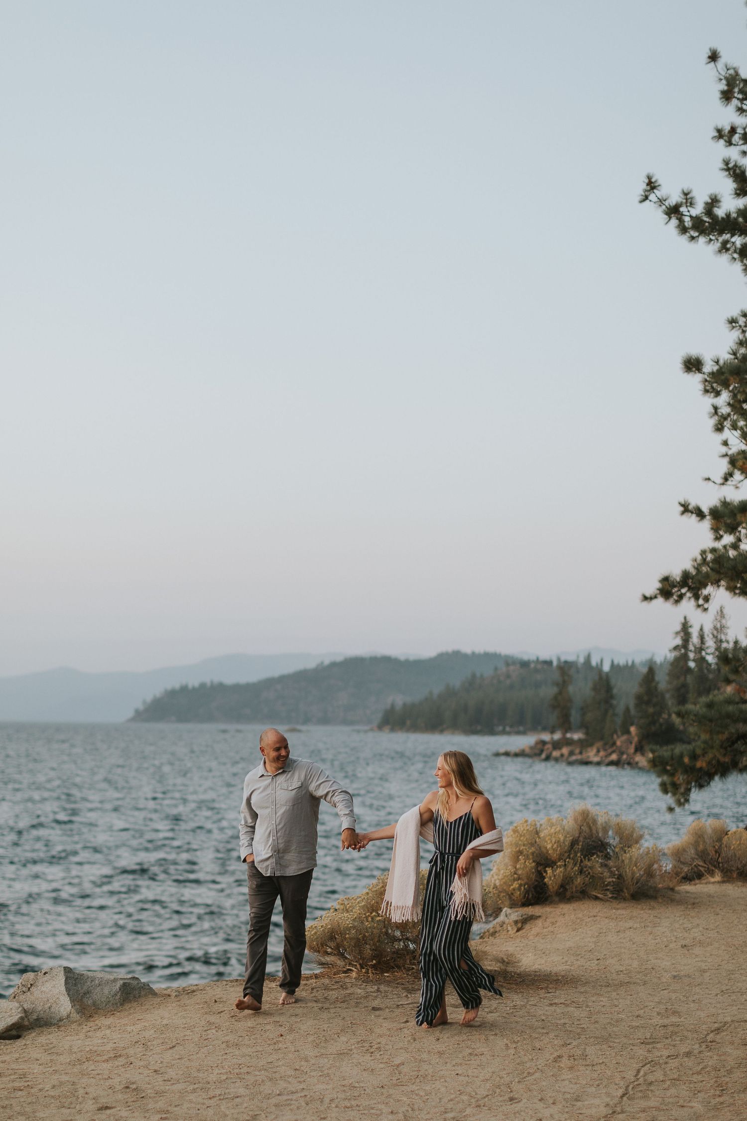Couple standing by a scenic lake with mountains and pine trees in the background.