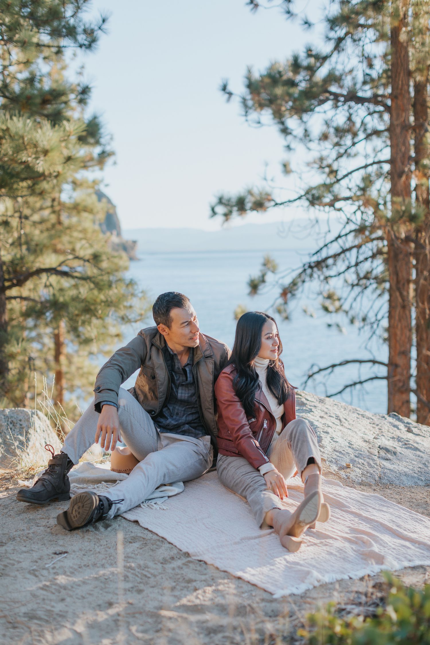 Couple relaxing on white blanket at sandy beach surrounded by pine trees.