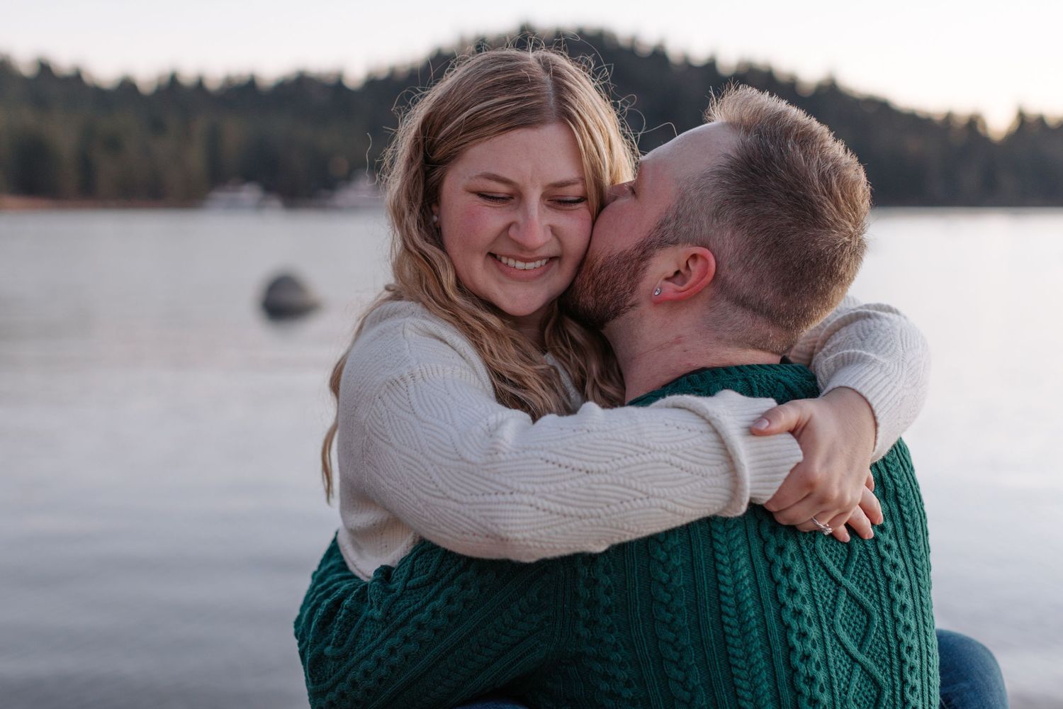 Couple embracing on beach with emerald green sweater and cream colored top against lake backdrop.