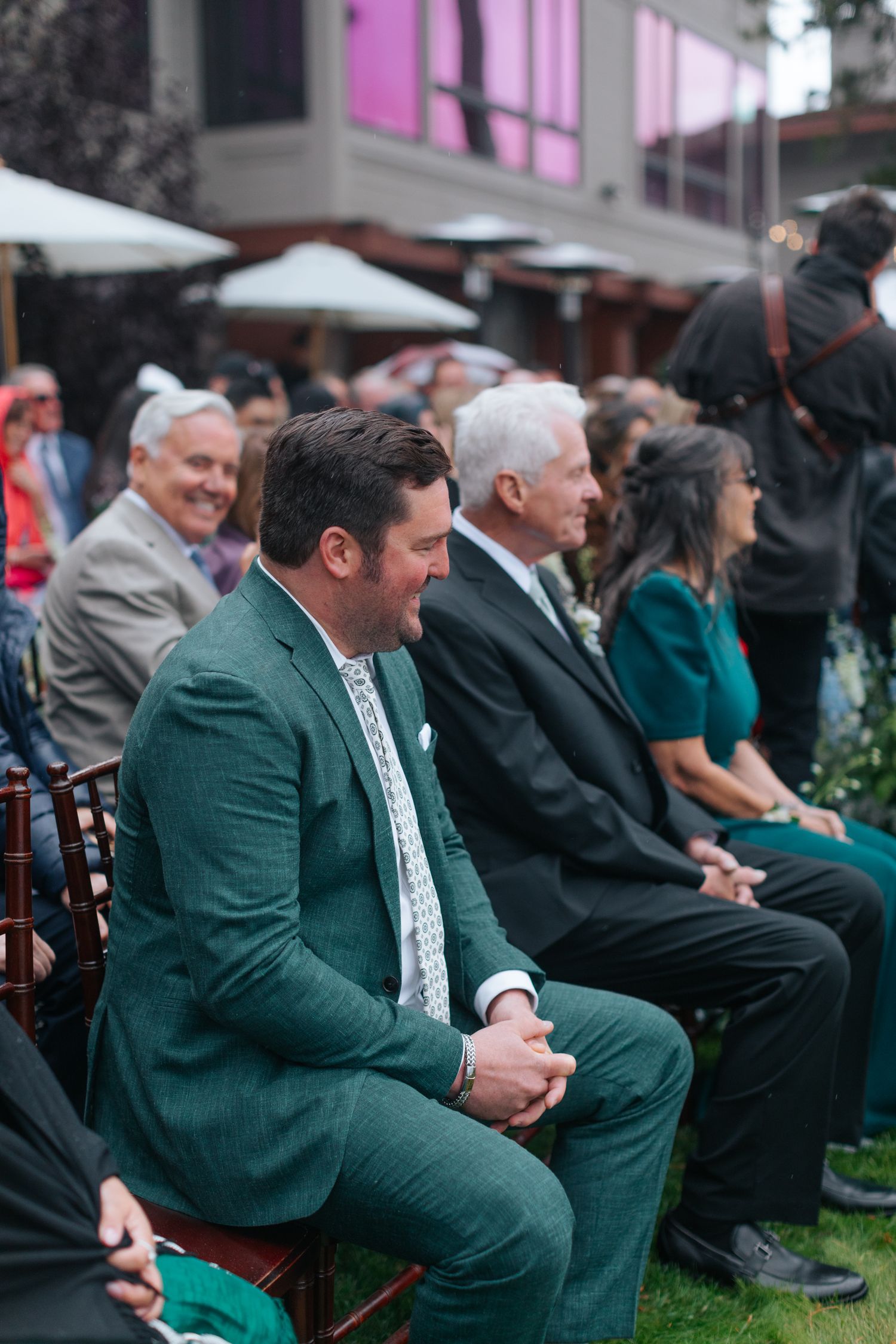 Wedding guests in formal attire seated in wooden chairs during outdoor ceremony.