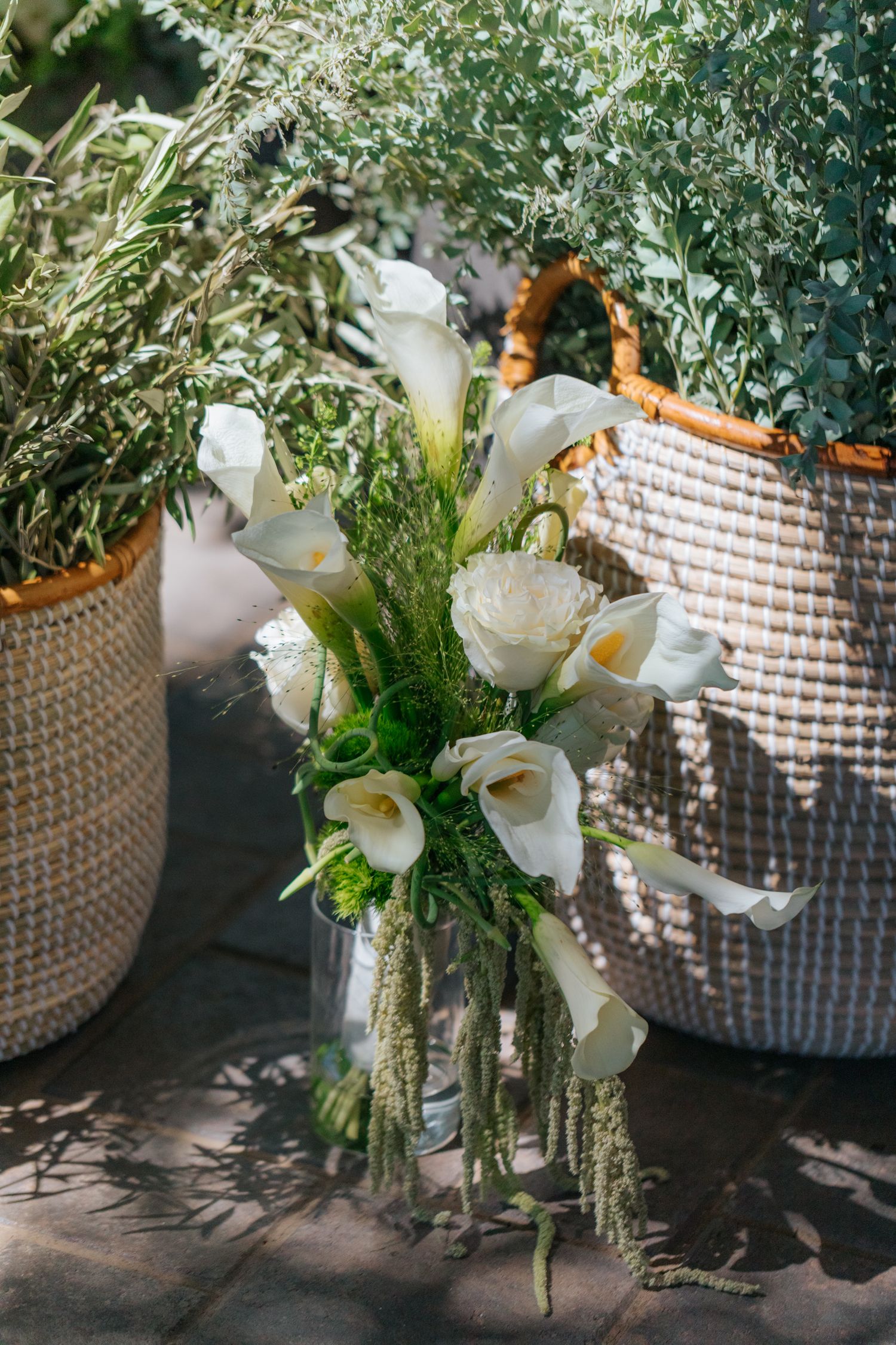 White calla lily and peony bouquet in glass vase beside woven baskets with greenery in sunlight.