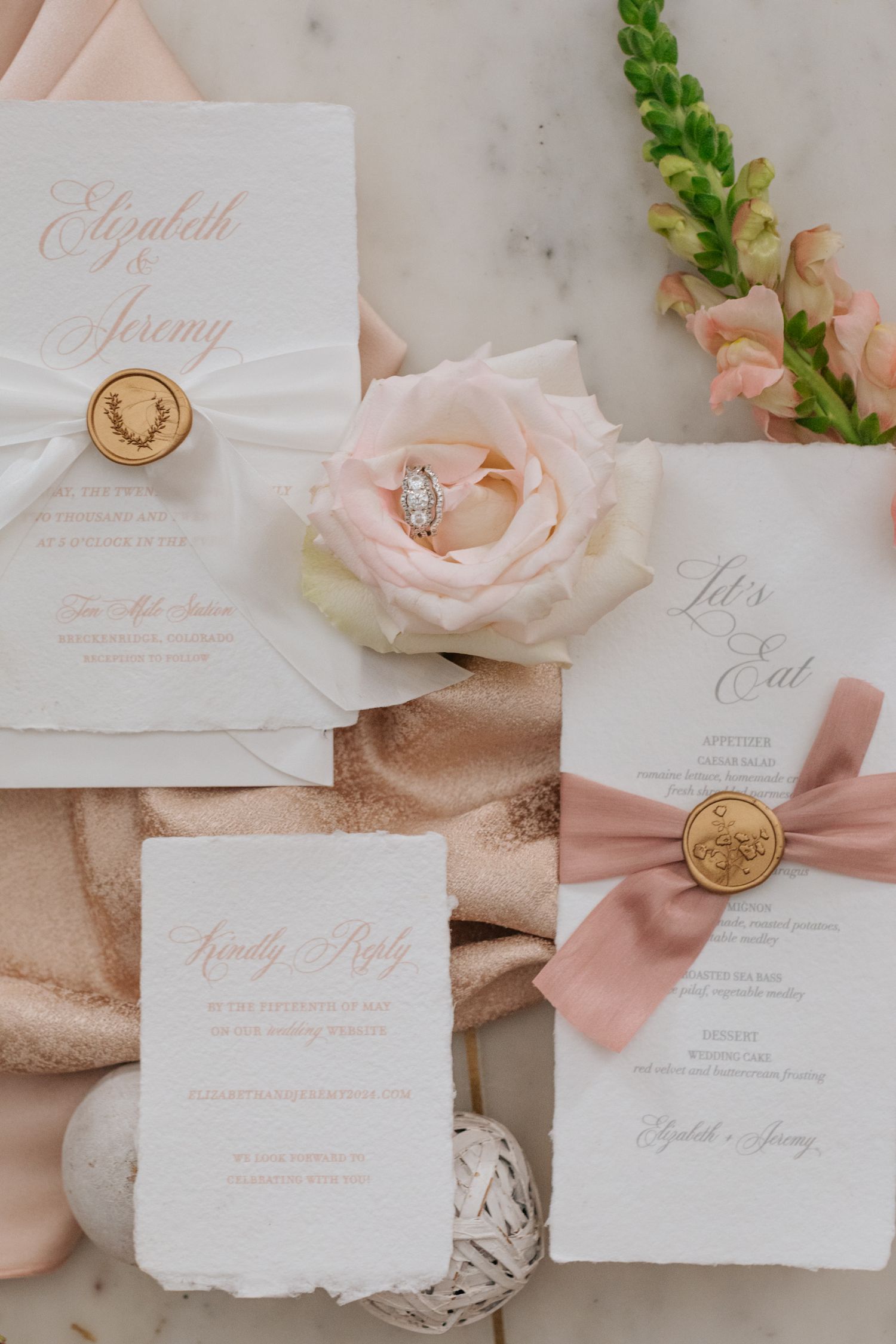 Elegant wedding stationery with blush pink ribbons, gold wax seals, and delicate roses on marble background.
