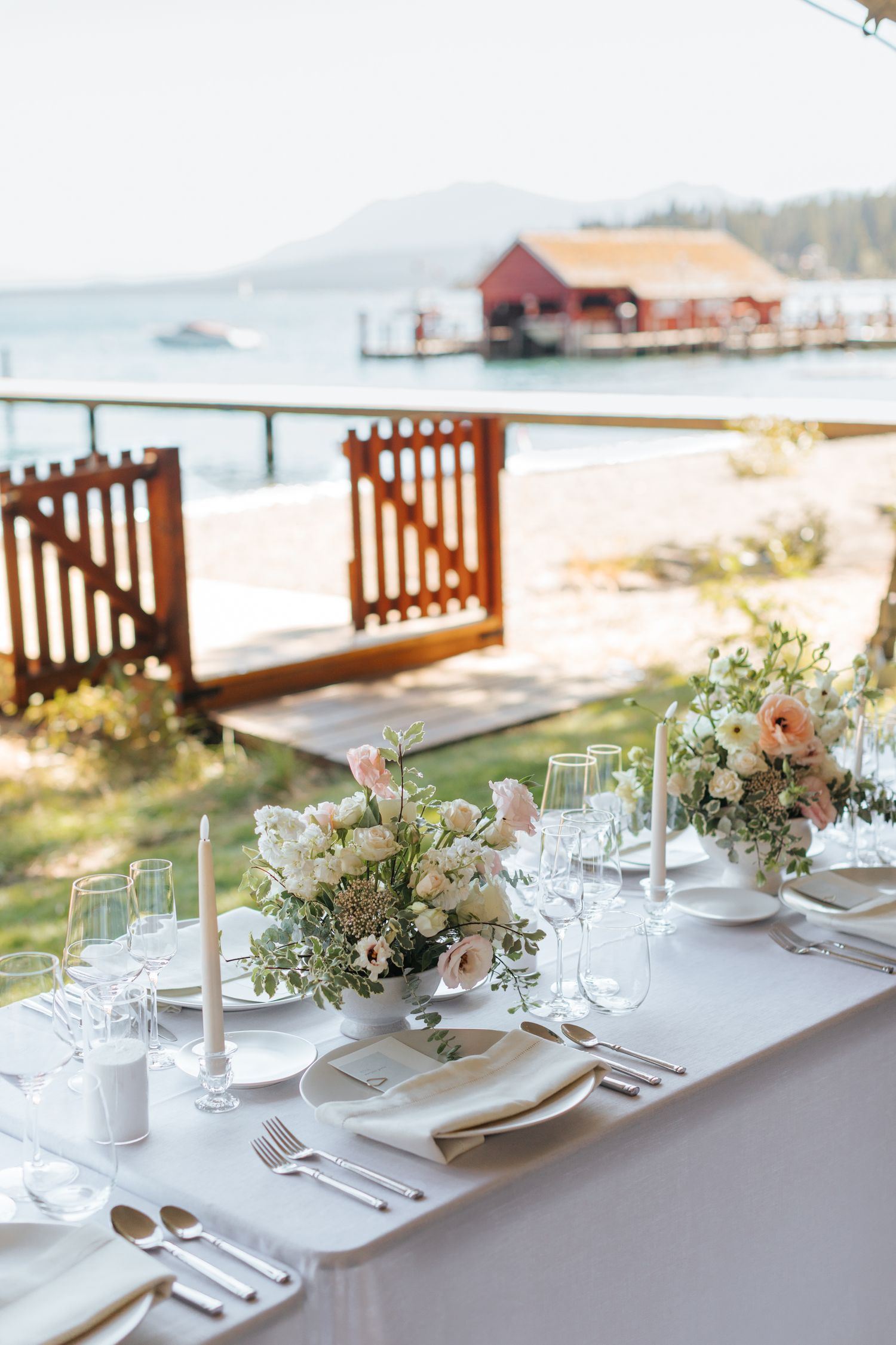Elegant lakeside wedding reception table with white linens, floral centerpieces, candles, and glassware overlooking a scenic waterfront.