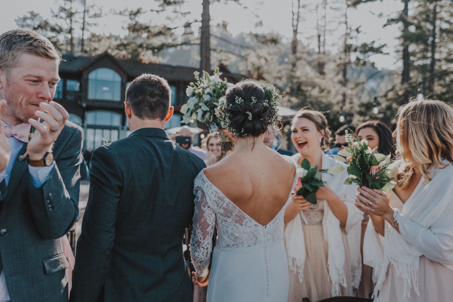 A joyful wedding celebration outdoors with guests and bridesmaids holding bouquets among pine trees.