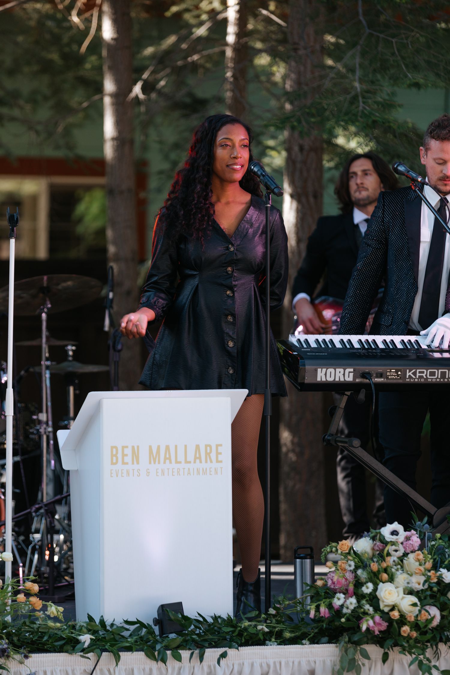Musicians perform behind white branded panels decorated with floral arrangements at an outdoor event.