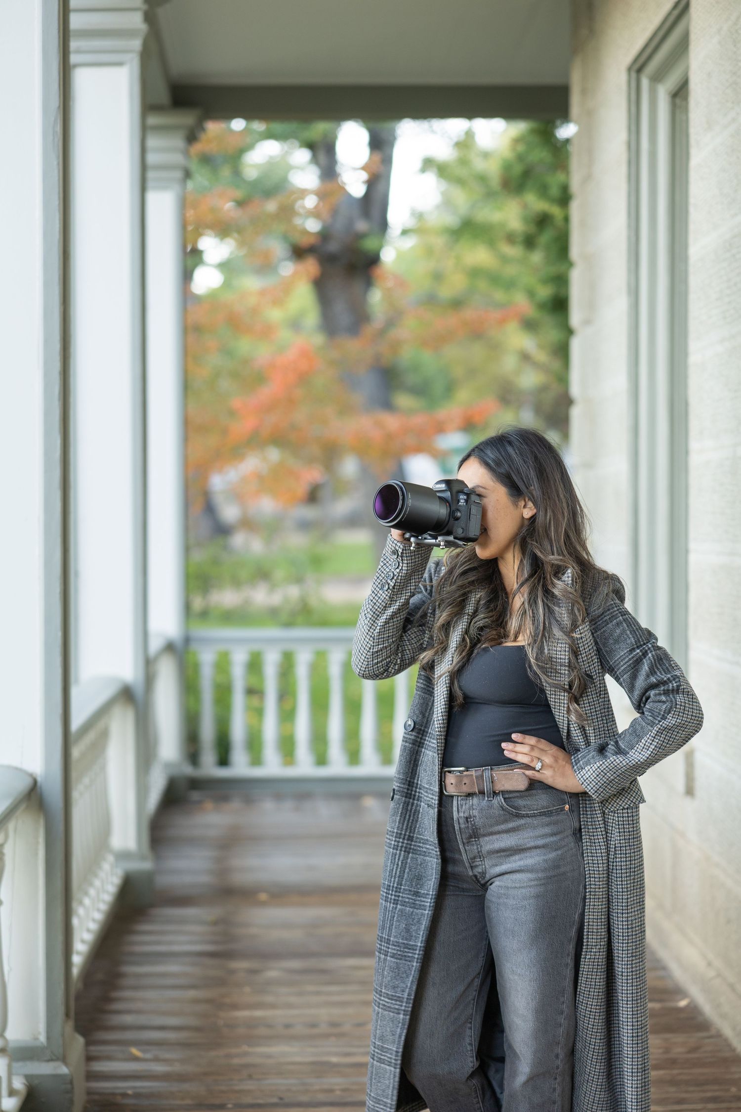 Photographer with long hair using professional camera on rustic porch with autumn scenery.