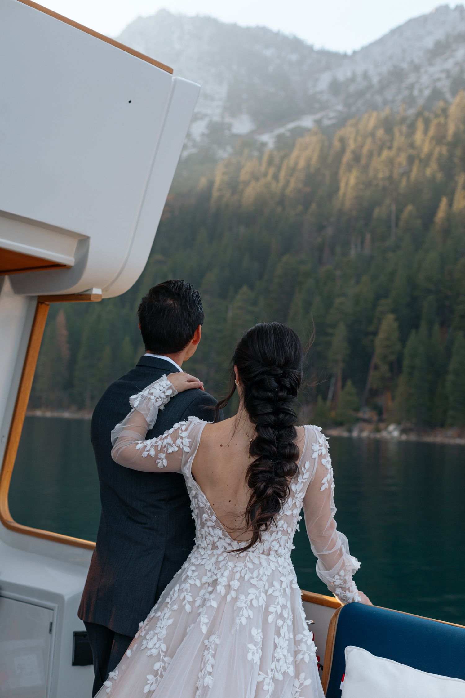 Couple on boat deck overlooking mountain lake with pine forest backdrop.