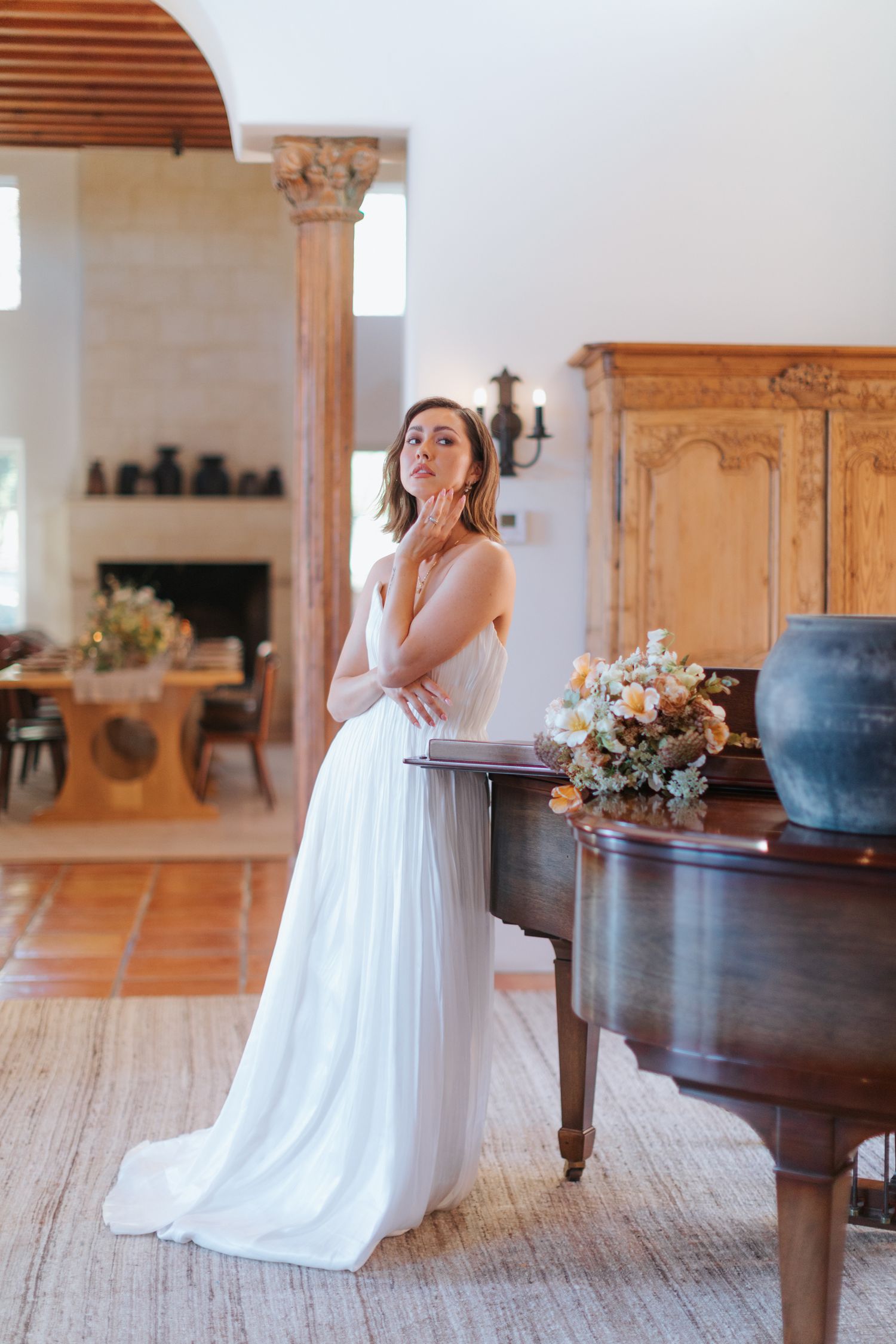 A bride in a flowing white gown poses elegantly near a grand piano in a light-filled room with floral arrangements.